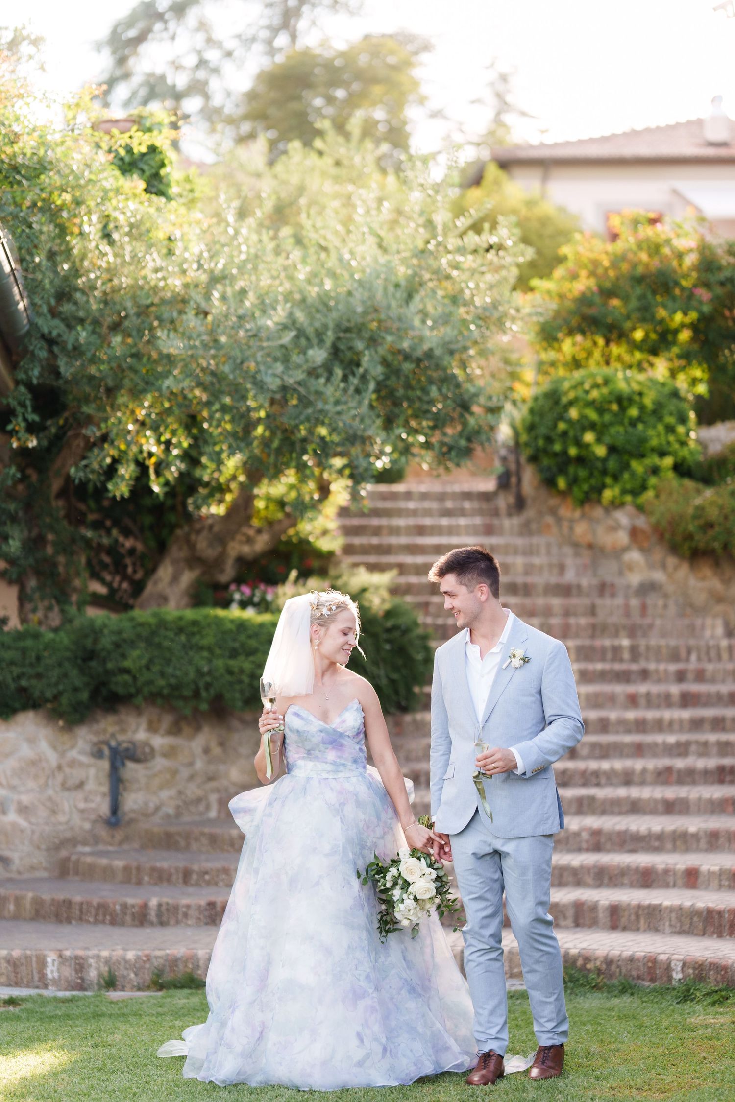 Bride in blue floral gown holds bouquet while groom in gray suit walks with her on green lawn with stone steps.