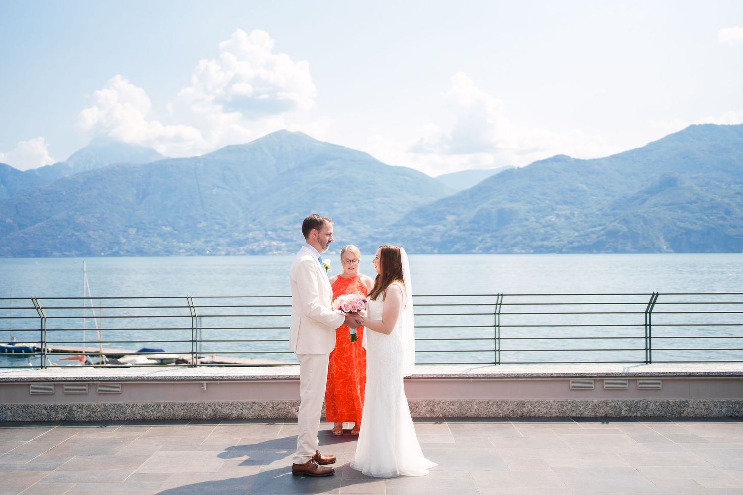 Couple exchanging vows with officiant on lakeside terrace with mountain backdrop at outdoor wedding ceremony.