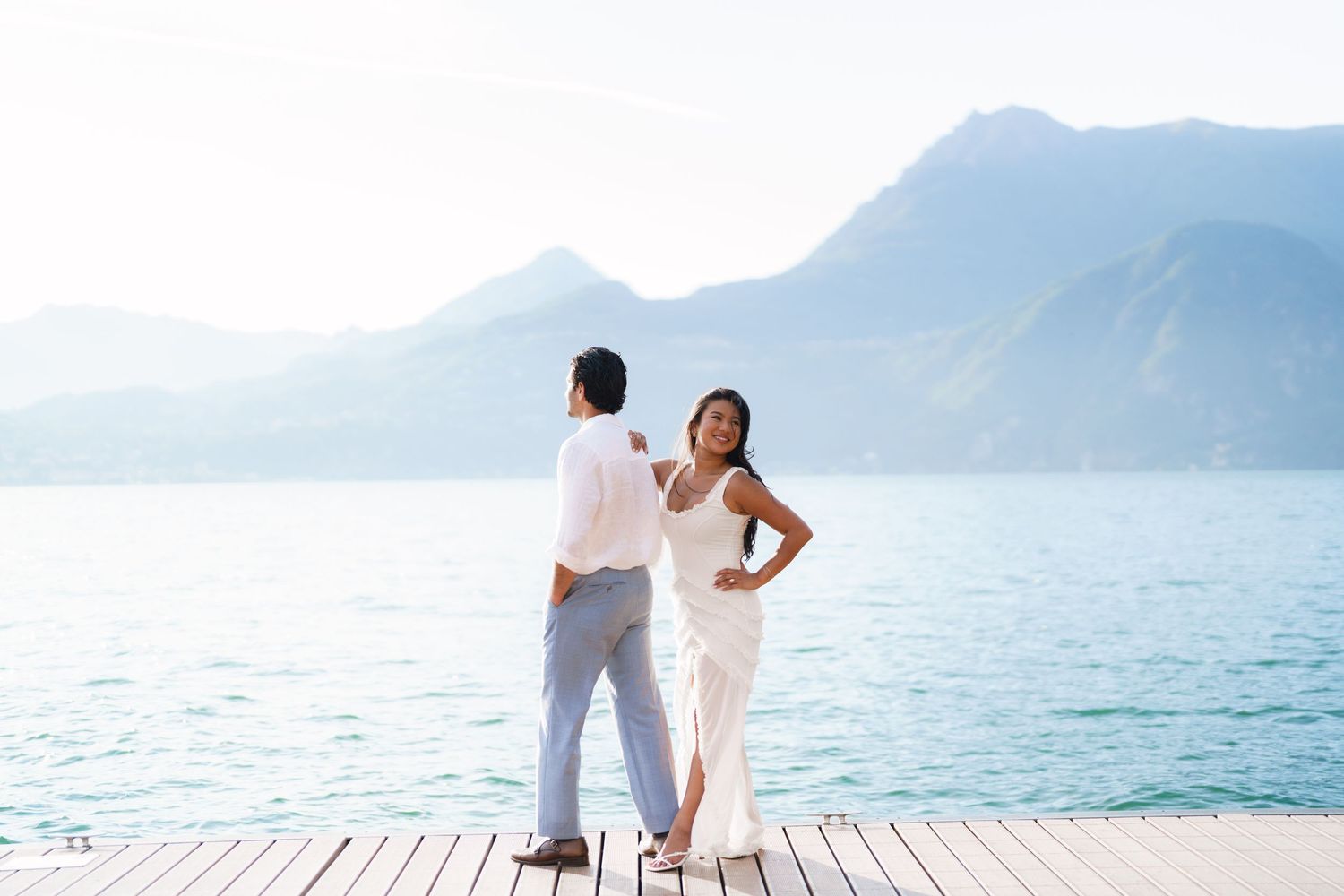 Couple in white outfits standing on a wooden dock by a turquoise lake with mountains in the background.
