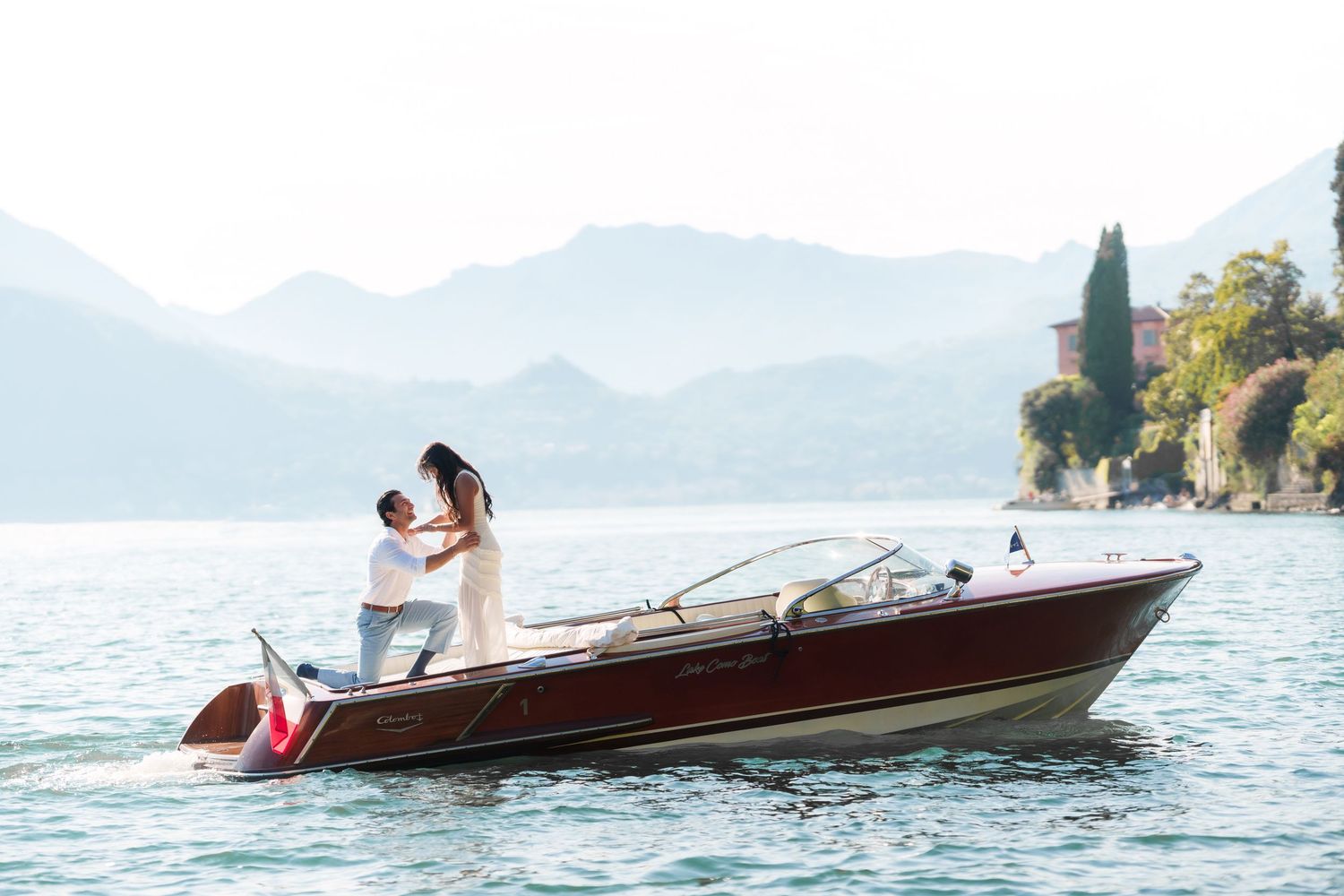 Couple embracing on a classic wooden boat on a serene lake with misty mountains and lush greenery in the background.