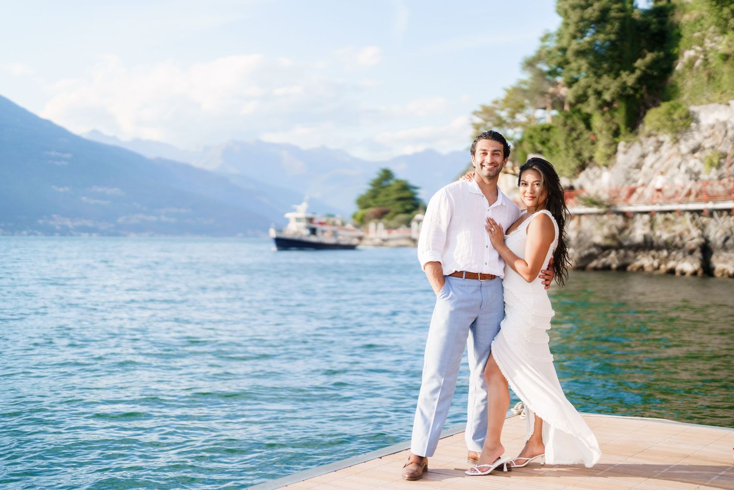 Couple posing on a lakeside dock with mountains and blue water in the background on a sunny day.