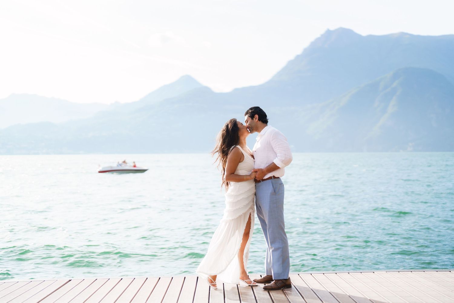 Couple sharing a romantic kiss on a lakeside dock with stunning mountain backdrop and turquoise water.