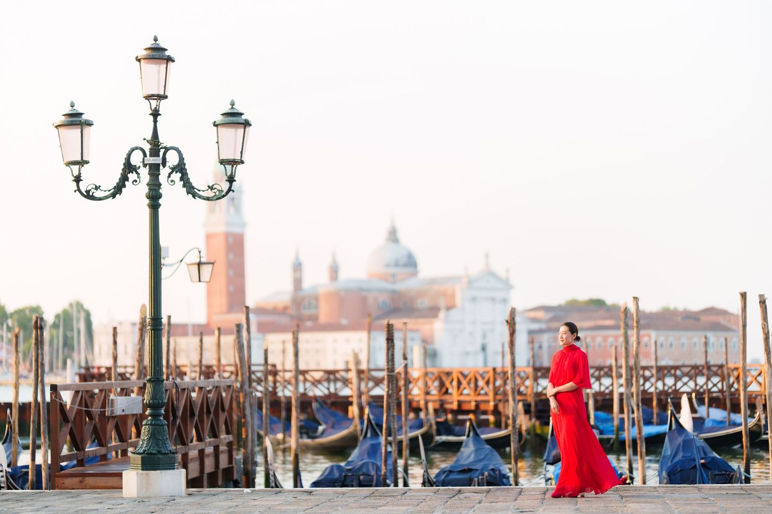 Woman in red dress walking near gondolas and lamp post in Venice, Italy with San Giorgio Maggiore in background.