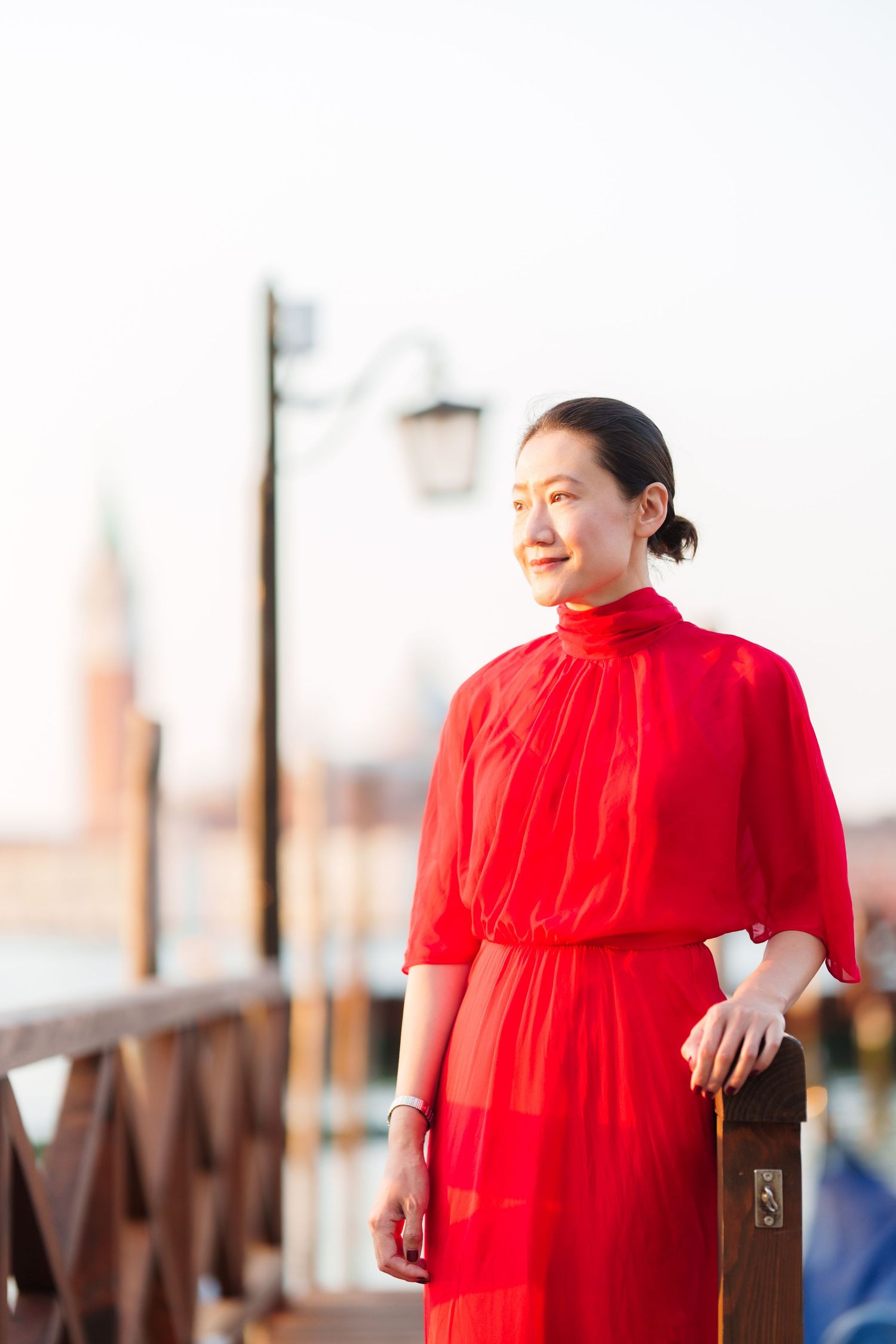 Woman in red dress standing on Venice waterfront with blurred campanile in background.