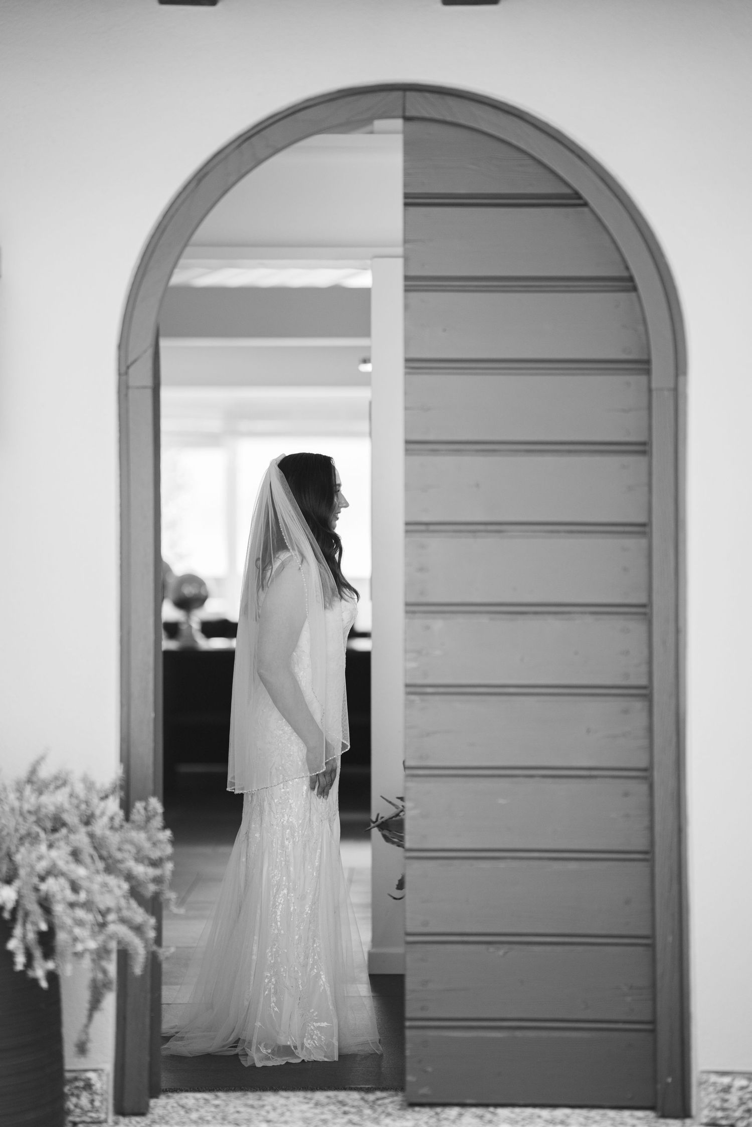 Black and white photo of a bride in a lace wedding dress and veil standing in an arched doorway.