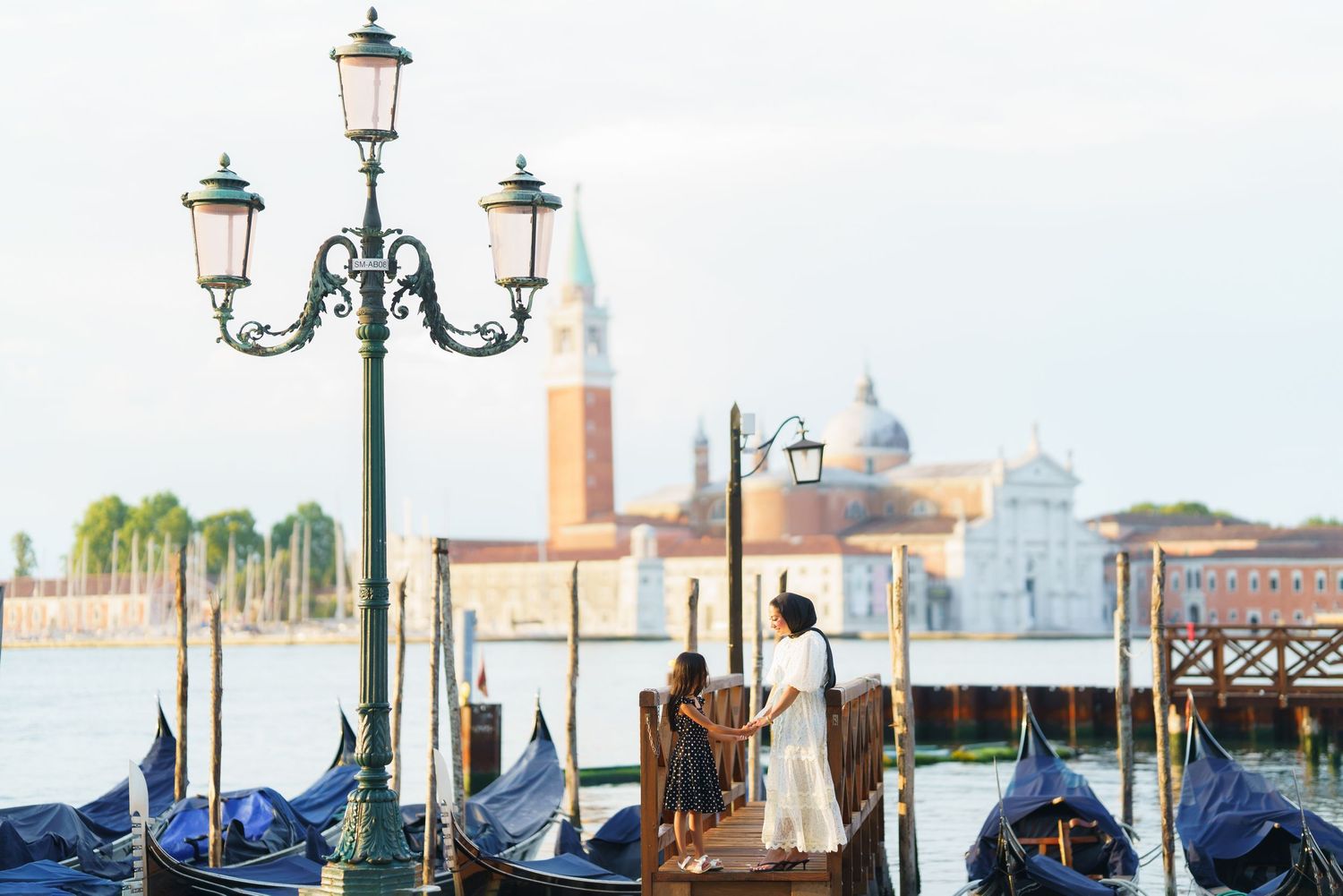 Couple on a Venice dock with gondolas and San Giorgio Maggiore church across the lagoon at sunset.