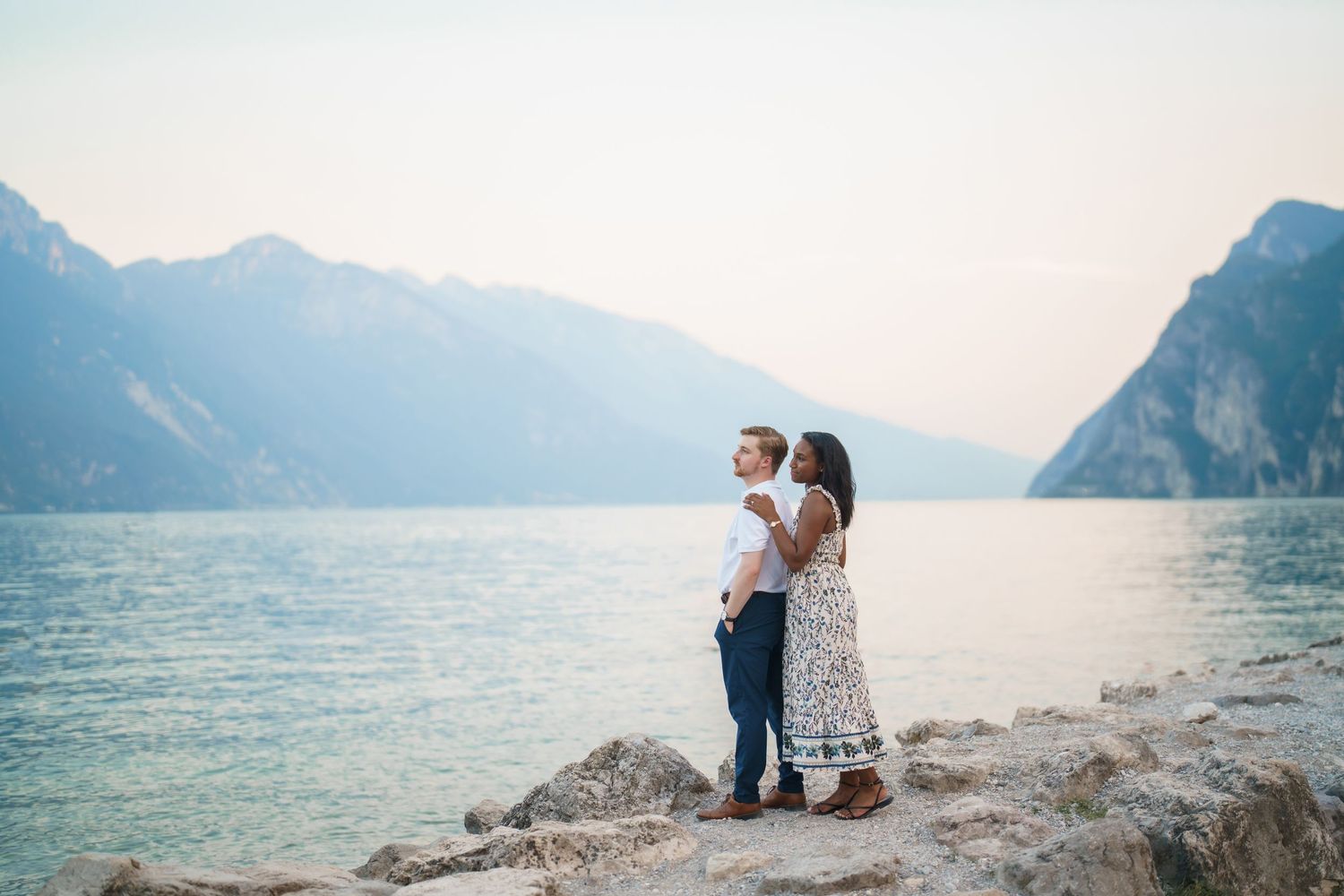 Romantic couple embracing by serene Lake Garda with dramatic mountain scenery at sunset.