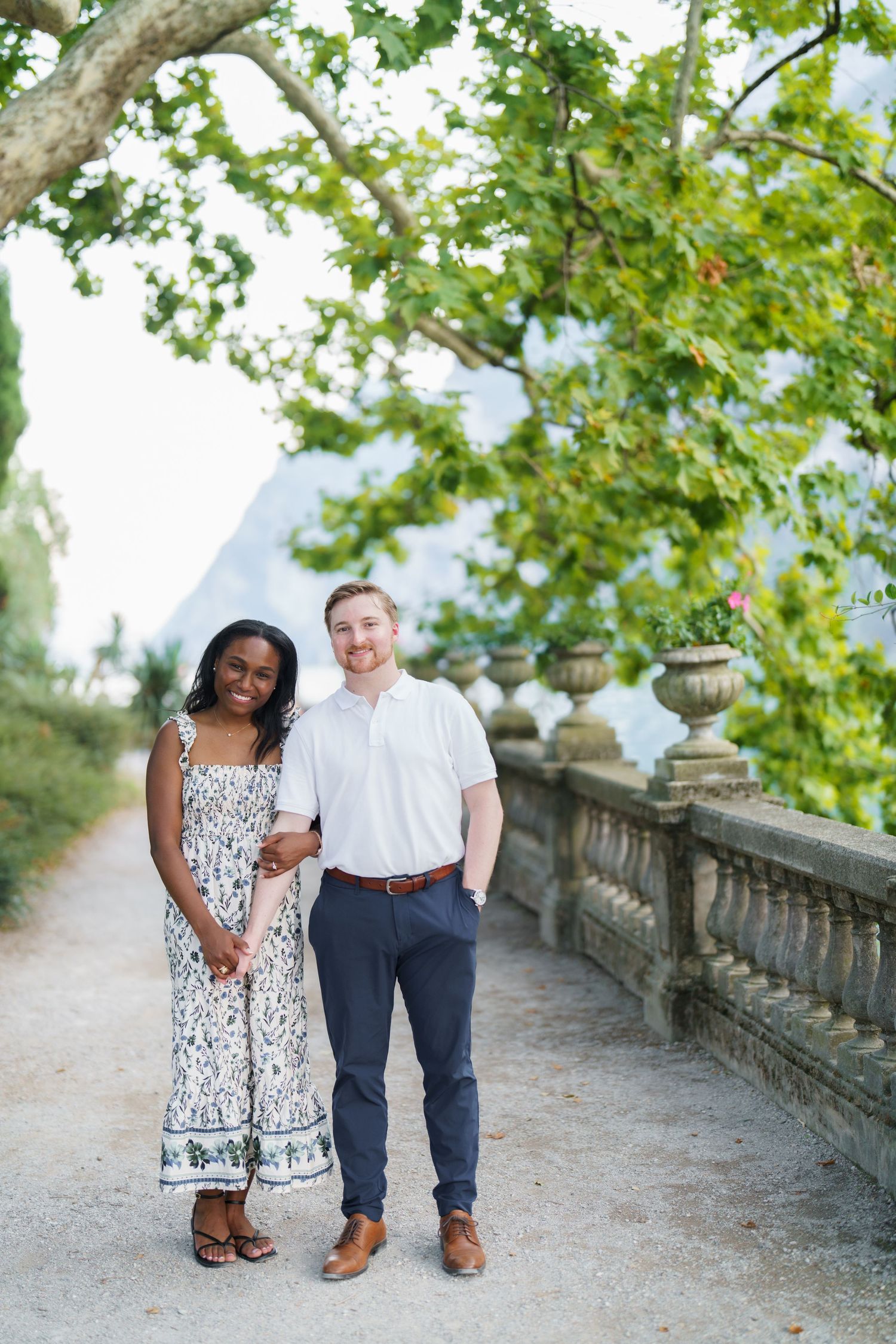Happy interracial couple posing on a garden path under green trees with stone railing backdrop.