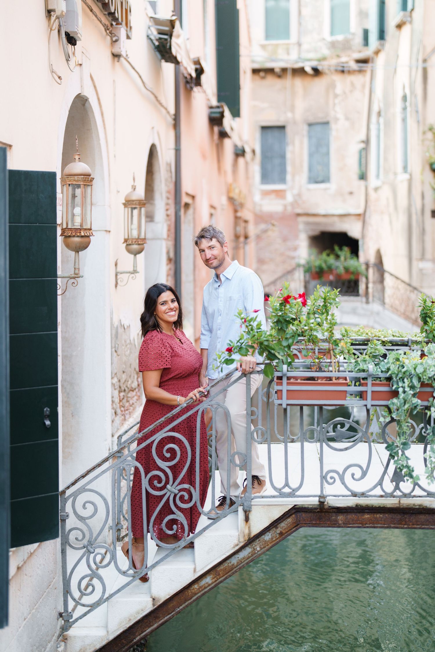 Couple posing on ornate iron bridge over Venice canal surrounded by historic buildings and red roses.