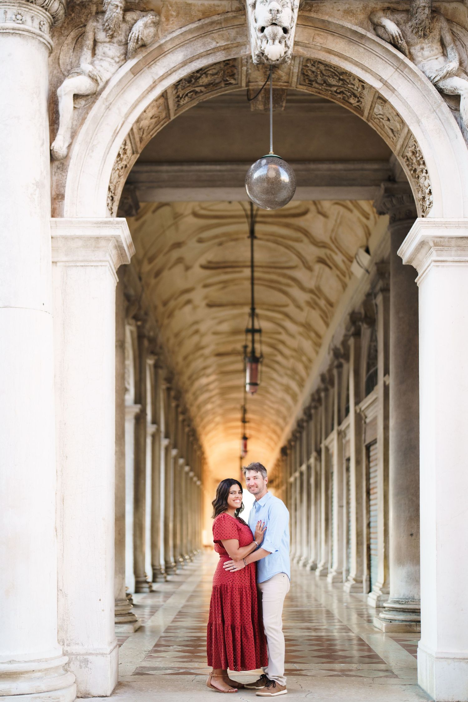 Couple embracing under ornate arched colonnade in Venice, woman in red dress, man in light blue shirt.