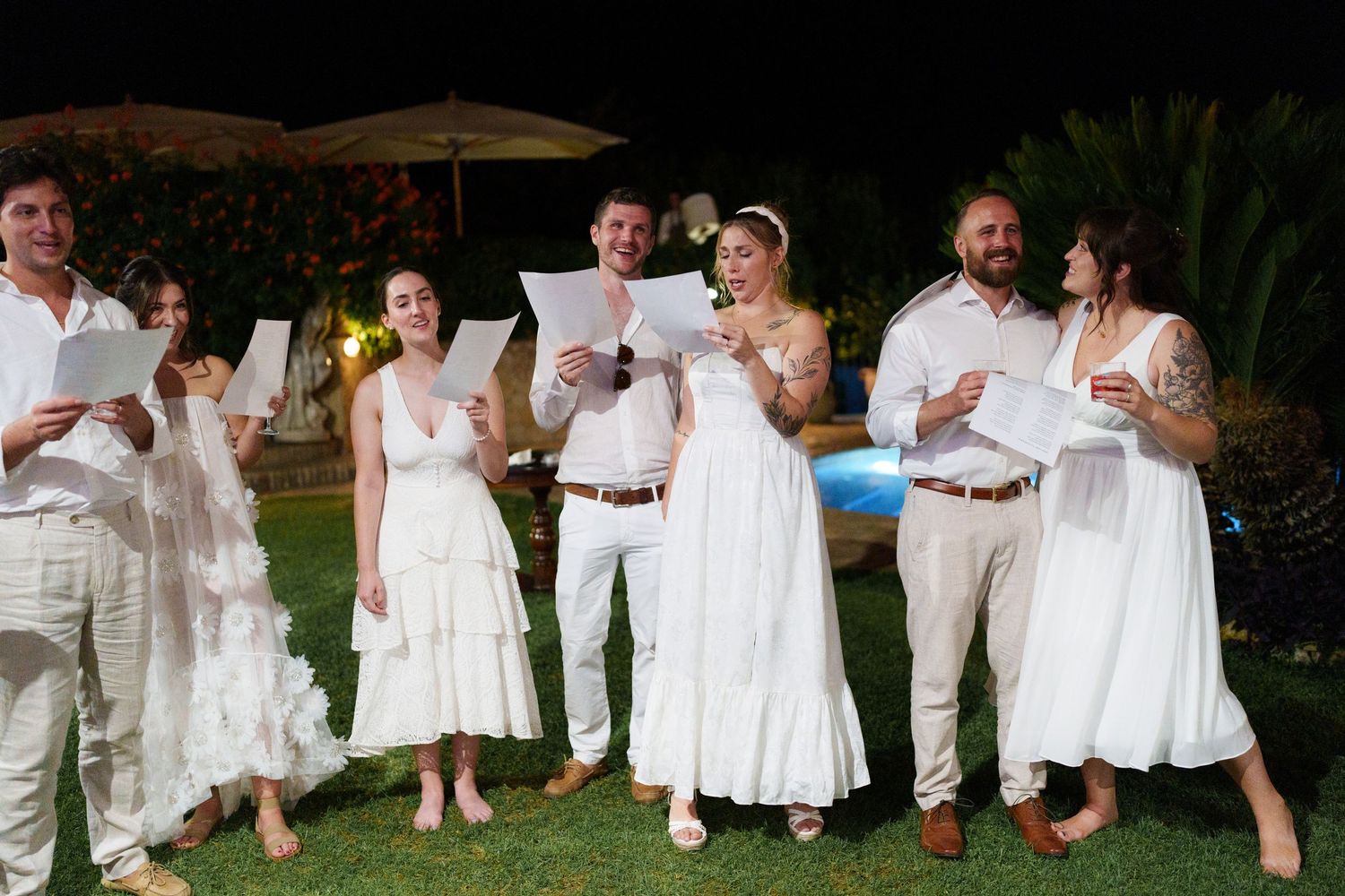 Group of people dressed in white holding papers at an outdoor evening wedding ceremony near a pool.