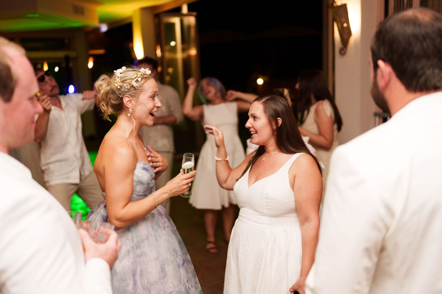 Two women in white dresses laugh and dance together at an elegant wedding reception with green ambient lighting.