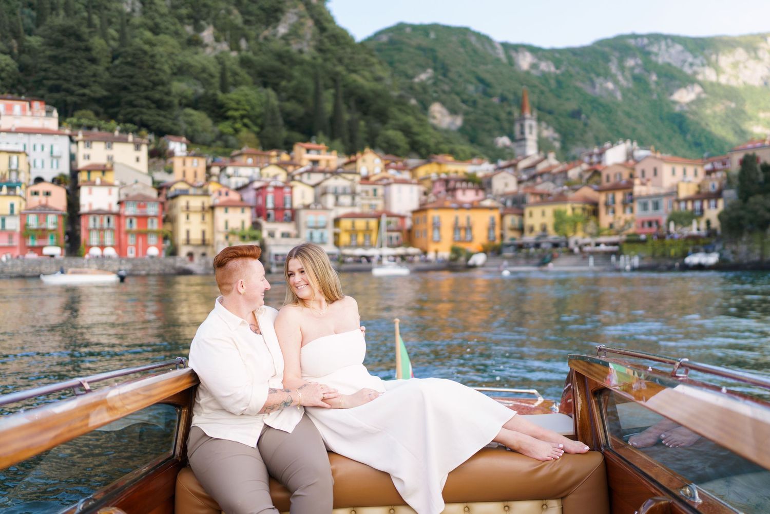 Couple sitting together on a wooden boat with colorful Italian village and lake in the background.