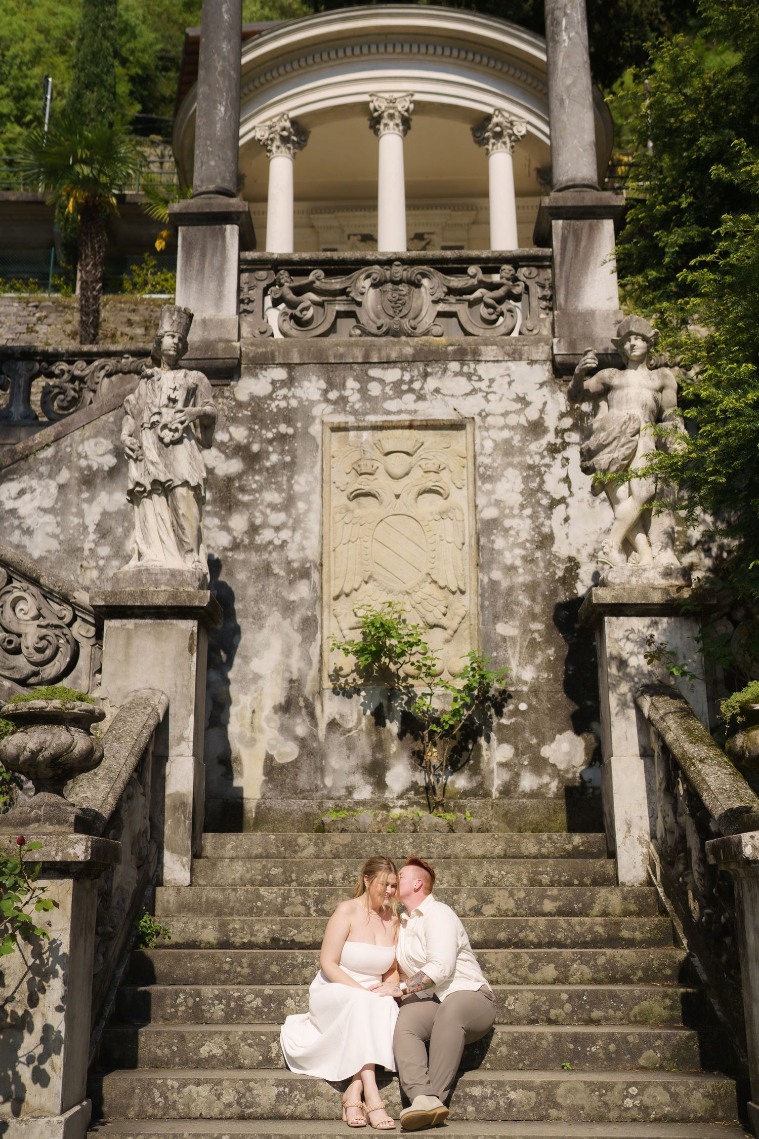 Couple sitting on ornate stone steps of a classical European villa with statues and columns in lush garden setting.
