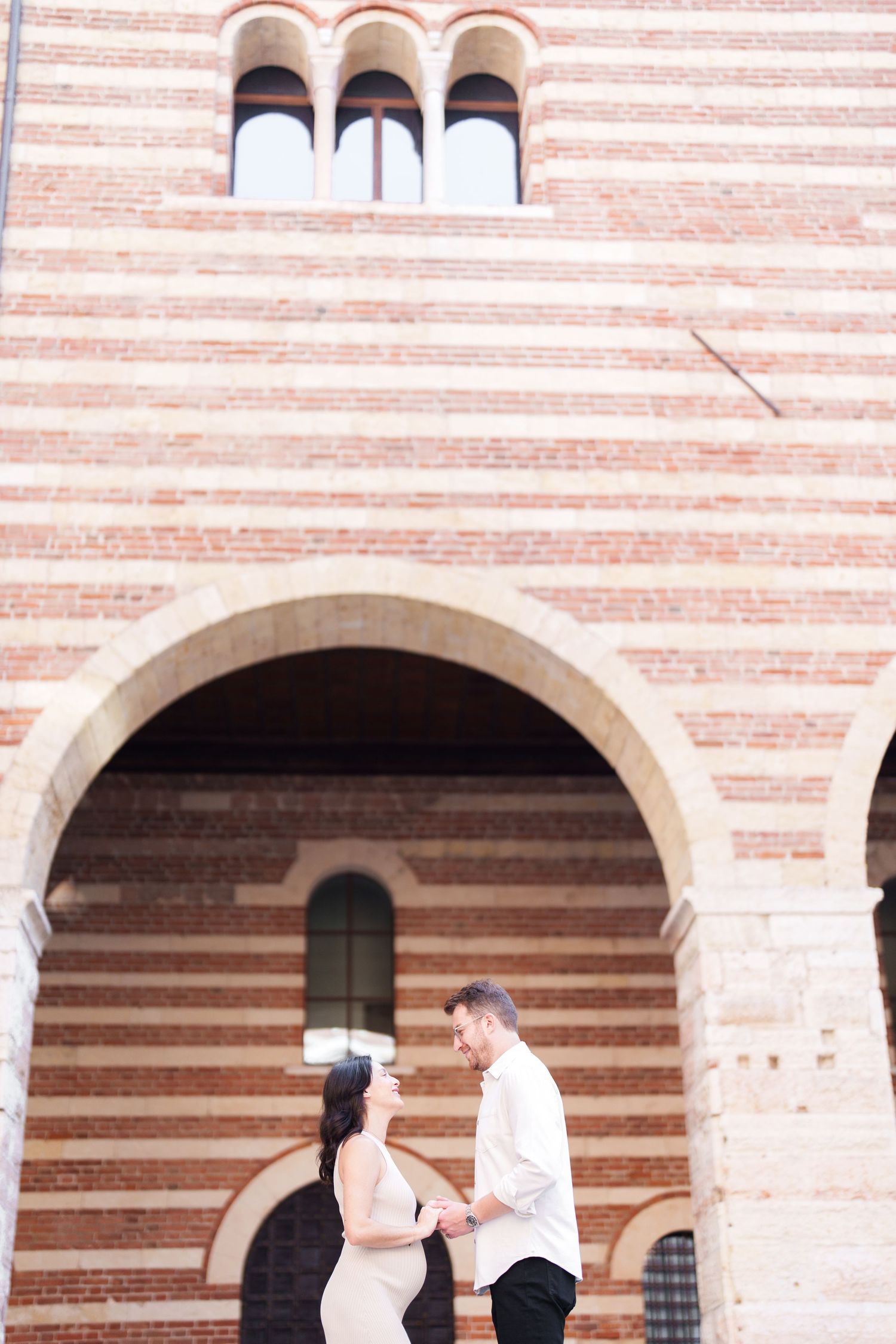 A couple stands together under a striped stone archway of a historic building with arched windows above.