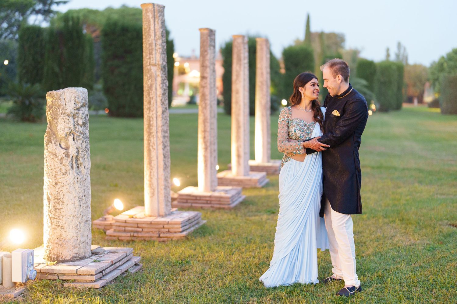 Couple poses among ancient stone columns in an outdoor garden setting.