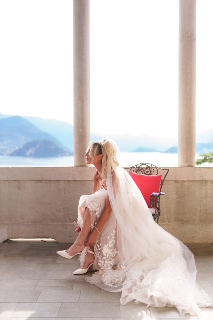 Bride adjusting shoe while seated on red chair under lakeside colonnade.