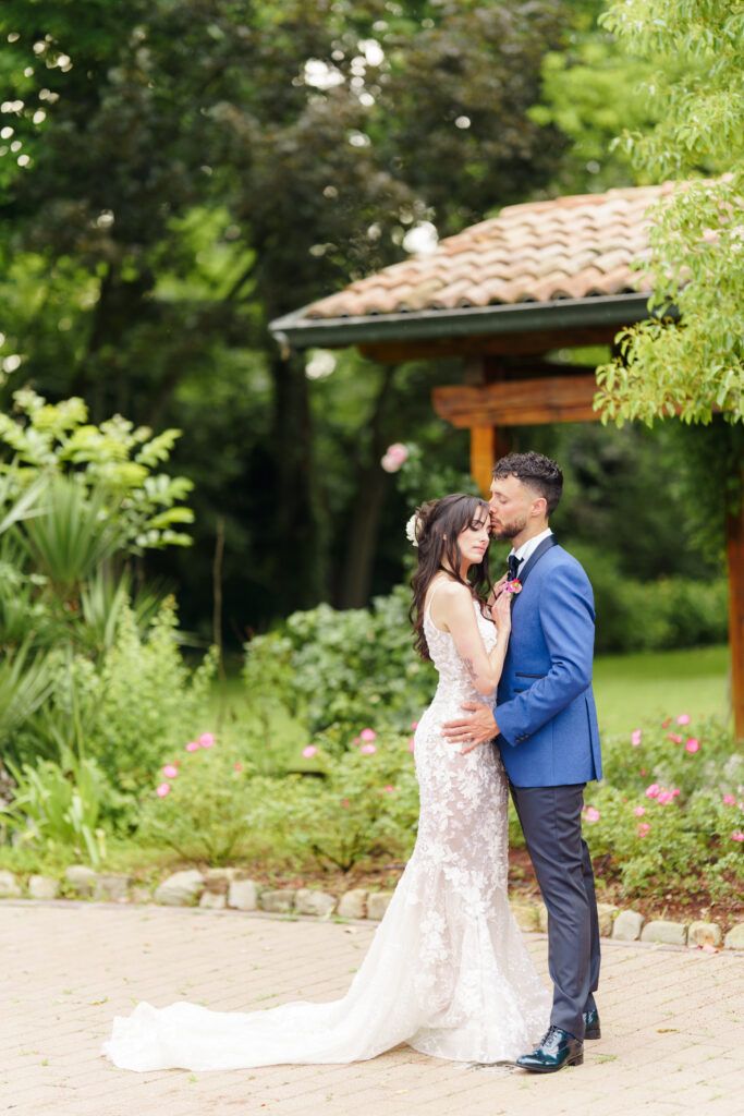 Romantic garden wedding photo shoot with couple embracing near a terracotta-roofed structure.