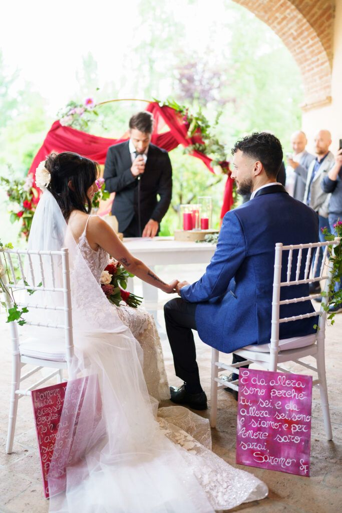 Wedding ceremony under brick archway with officiant speaking as couple sits on white chairs.