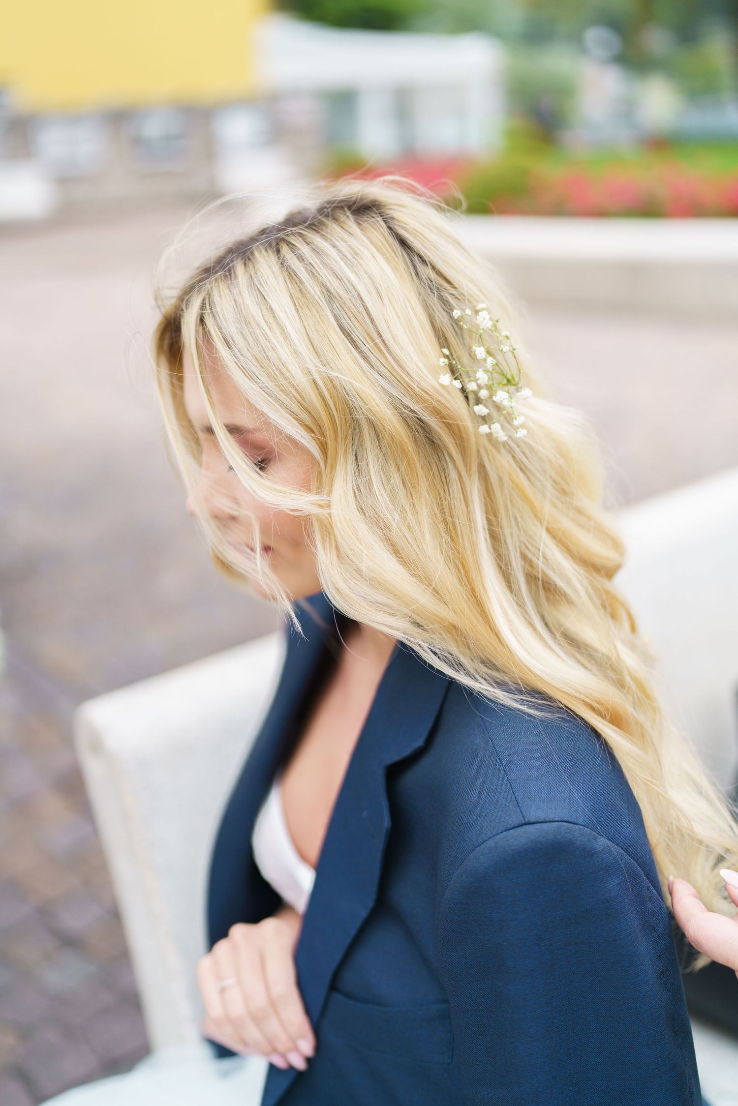 A person with long blonde hair wearing a navy blazer has small white flowers delicately woven into their hair.