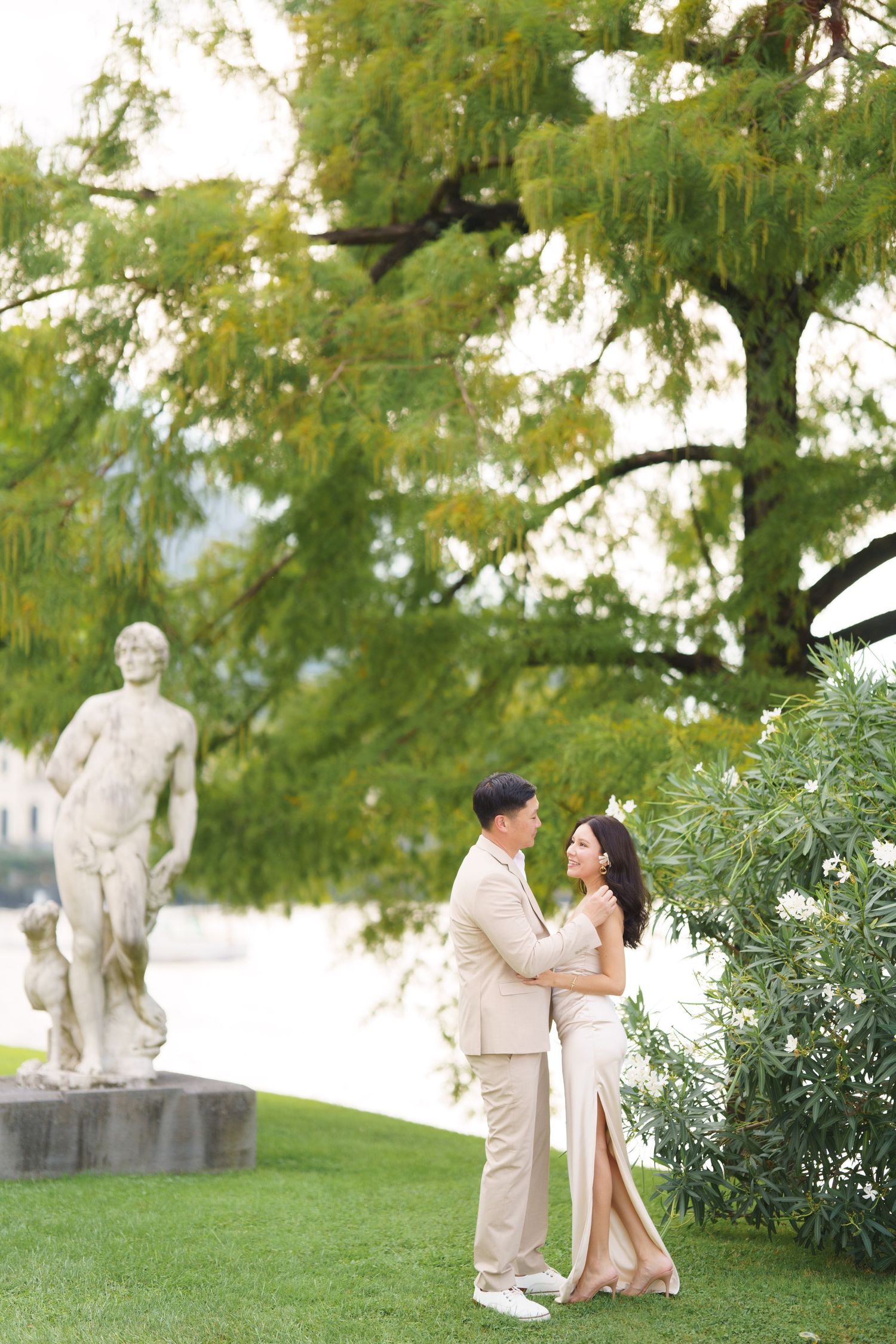 A couple embraces in an elegant garden setting with a classical stone statue and lush trees in the background.