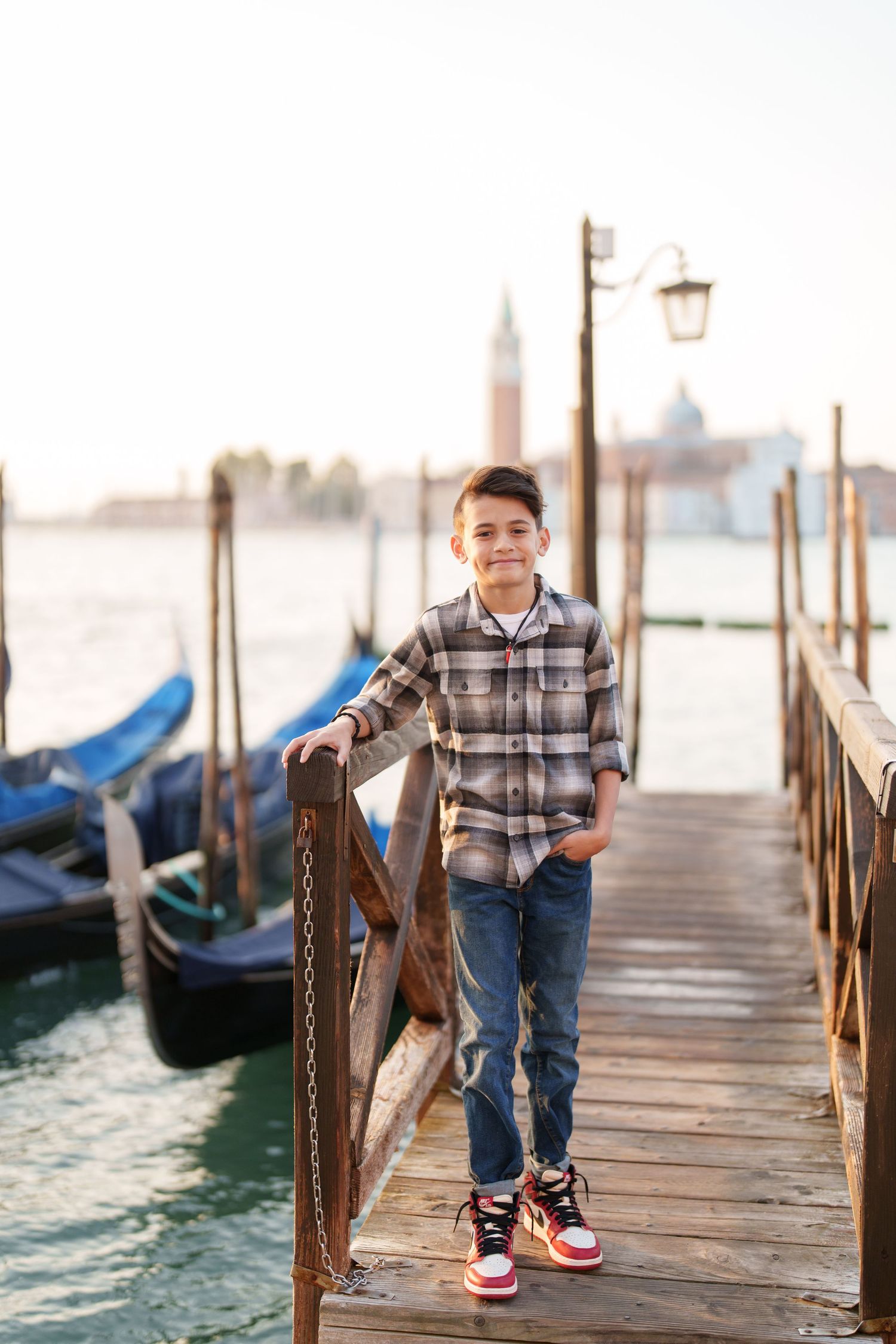 A young person stands on a wooden pier overlooking gondolas in Venice, wearing plaid and red sneakers.