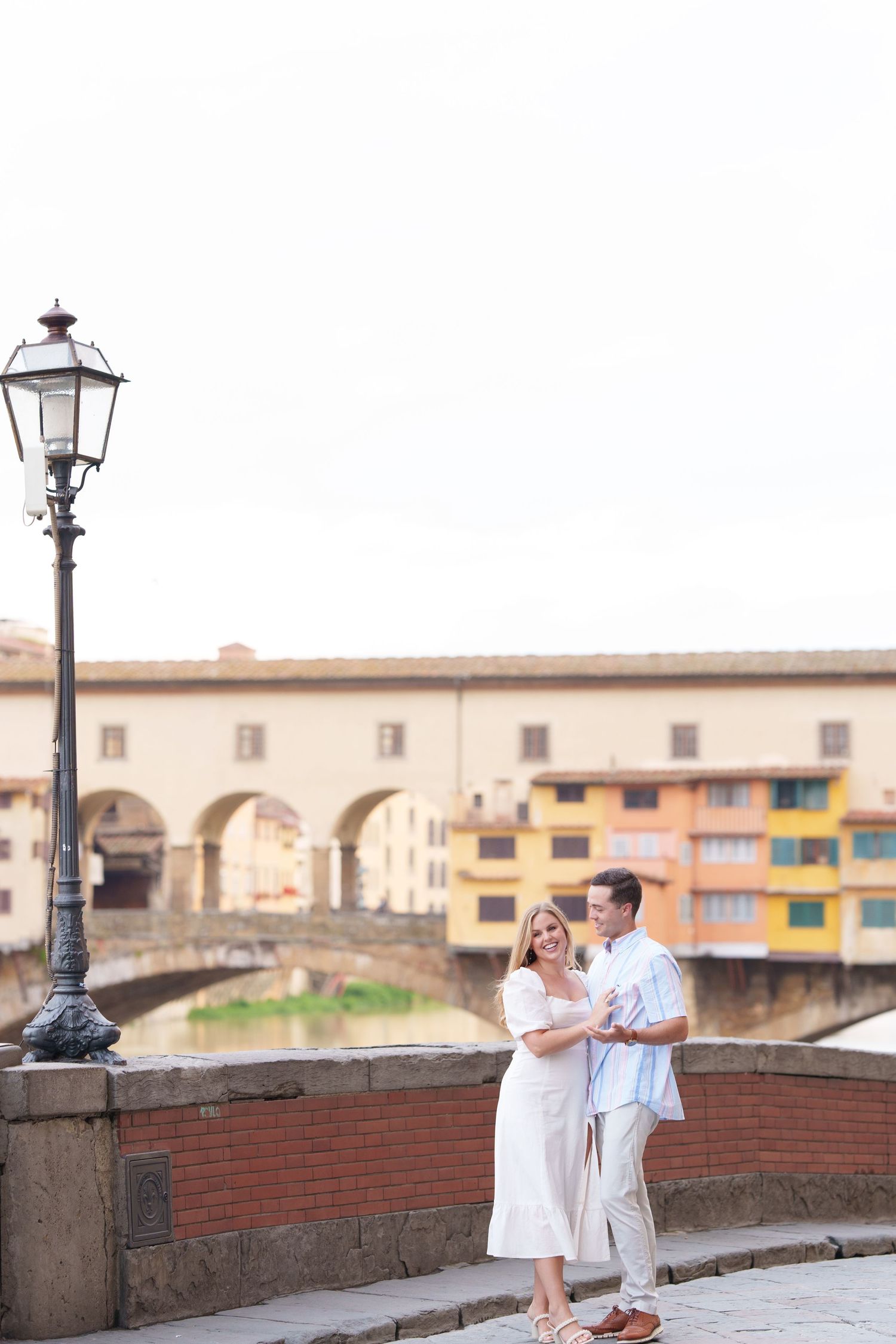 A couple in white outfits strolls along a scenic brick wall overlooking Florence, Italy during a romantic photoshoot.