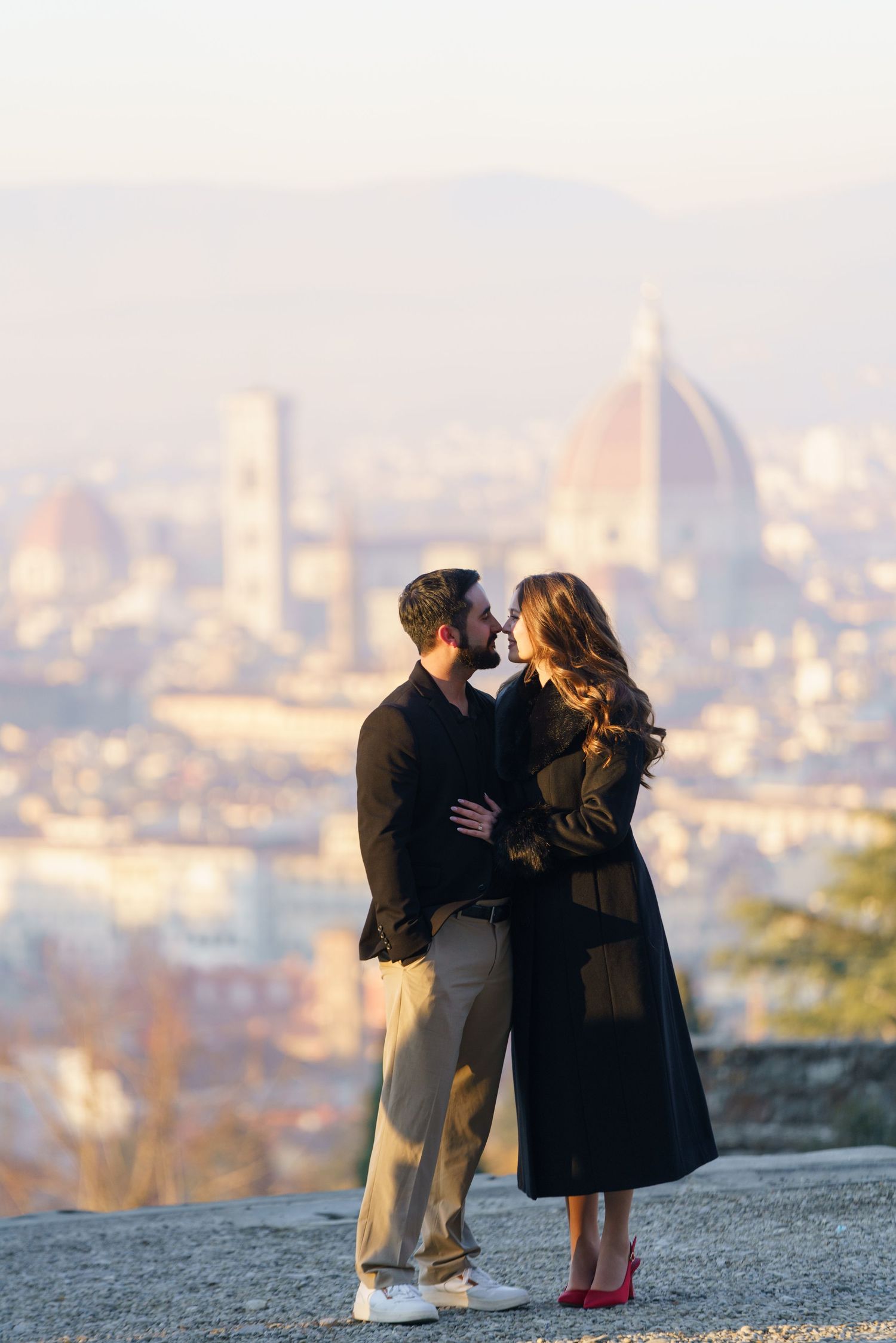 Couple embraces on hilltop overlooking Florence Italy with iconic Duomo cathedral visible in misty cityscape background.
