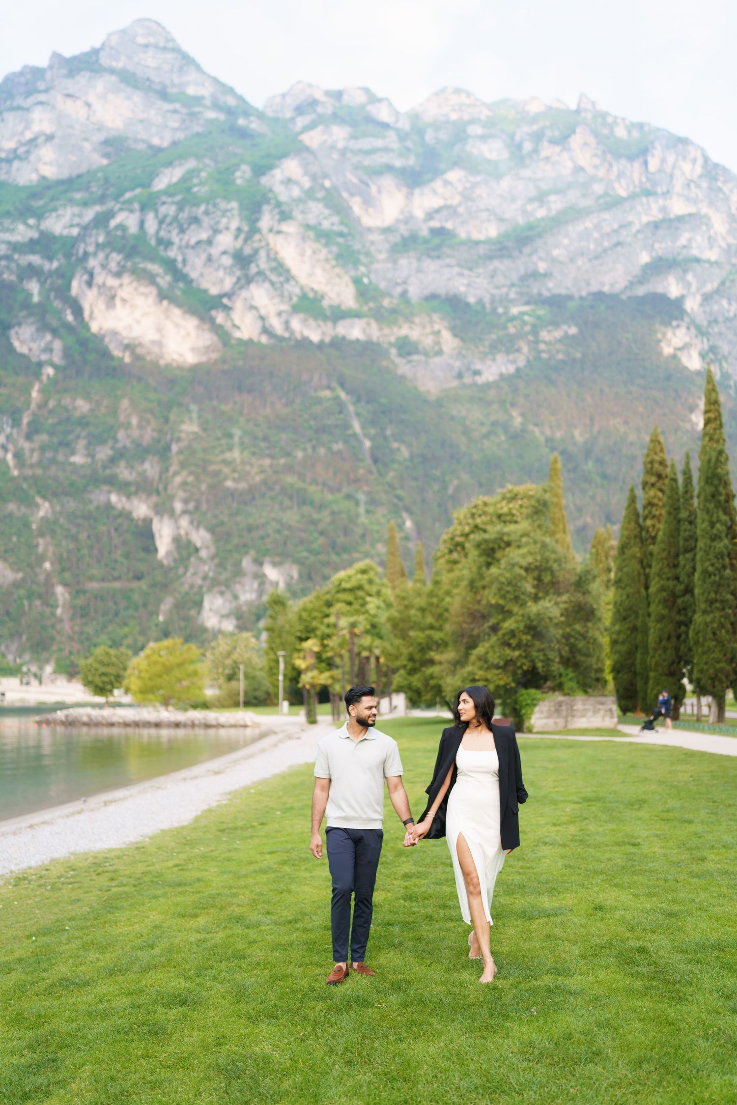 A couple walks hand in hand on green grass along a scenic mountain lake in the Alps.