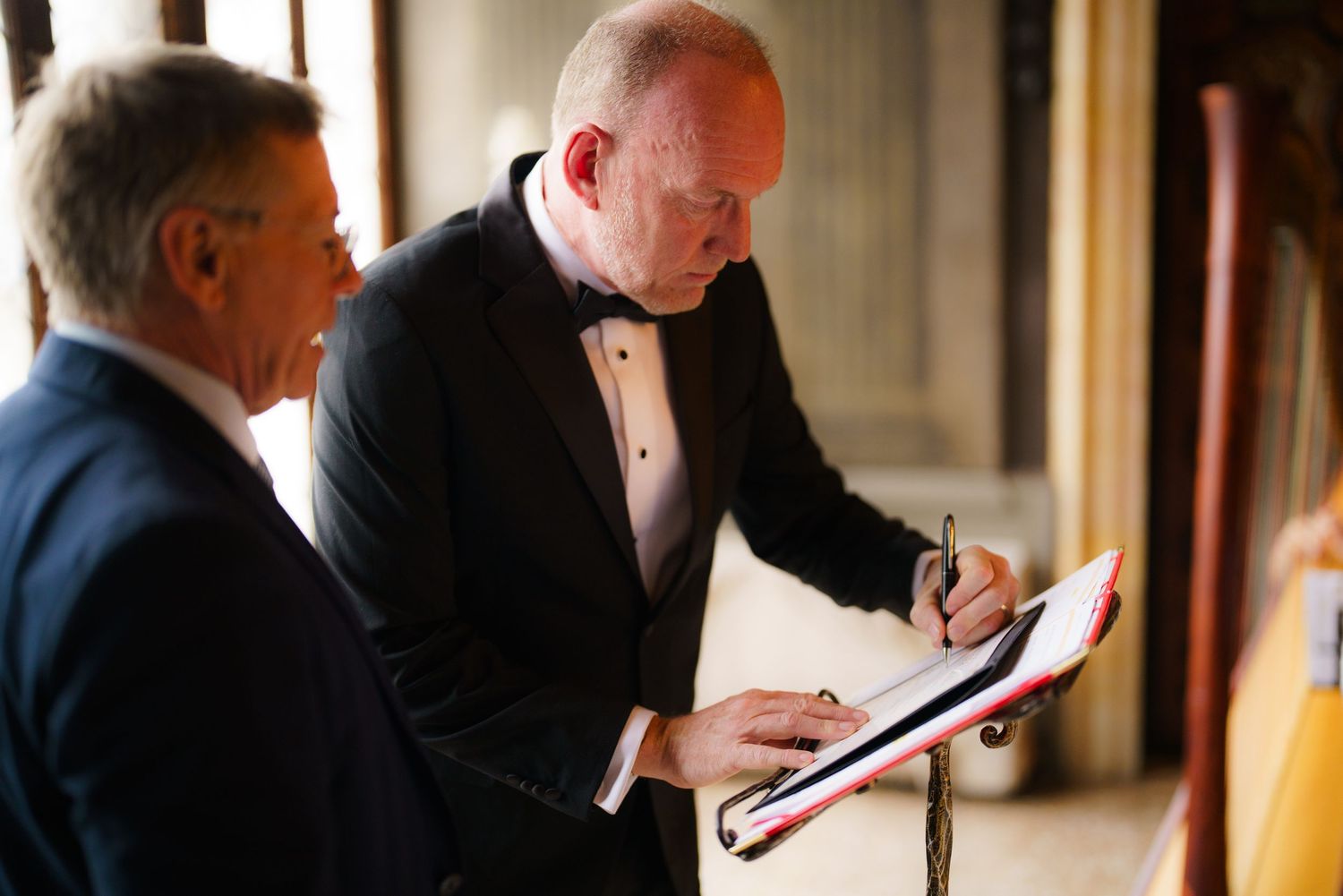 Two men in formal attire stand in a hallway while one signs a document on a clipboard.
