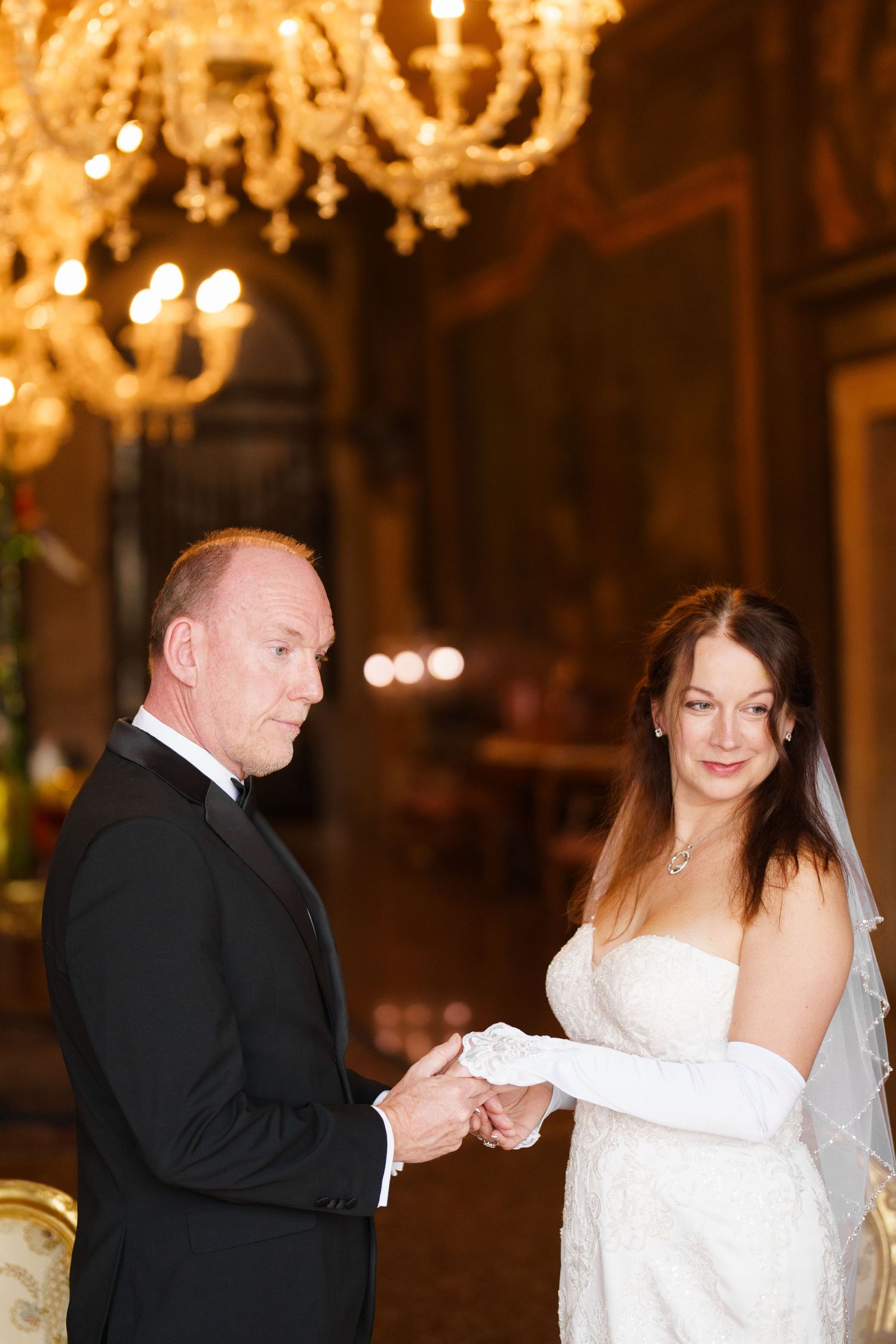 A couple shares a romantic dance moment in an elegant ballroom with chandeliers overhead.