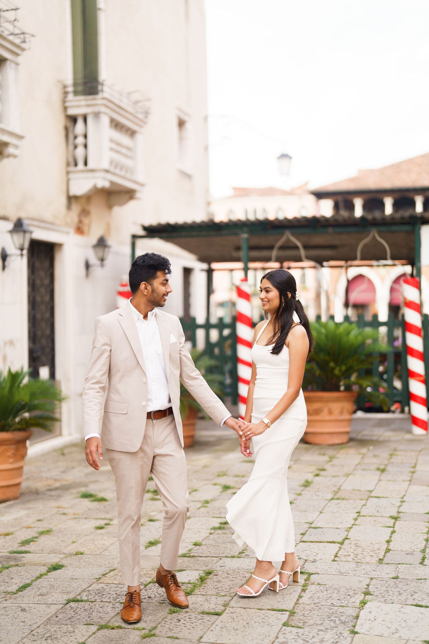 A couple in white wedding attire dances playfully on a stone patio with red and white striped poles in the background.