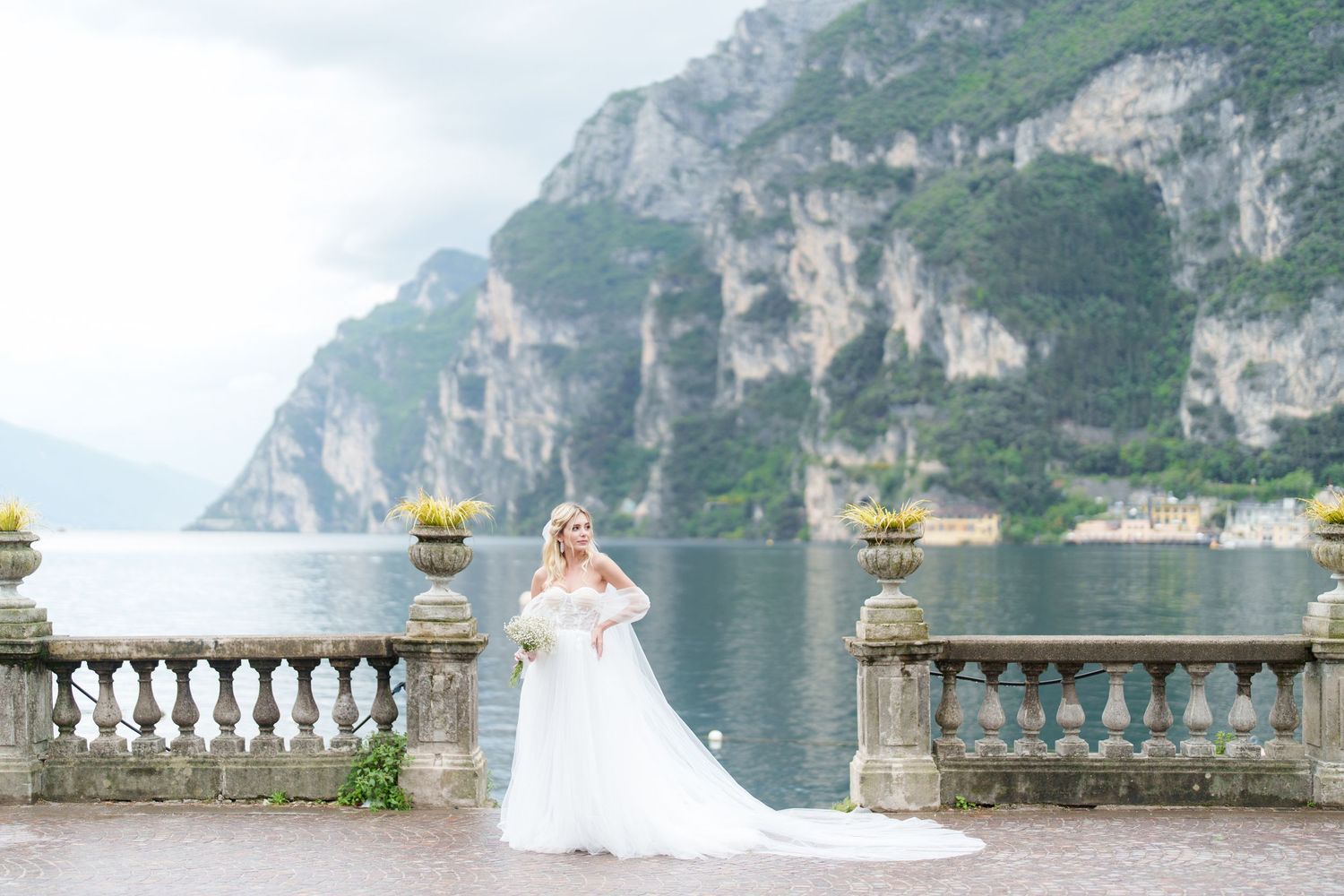 Bride in white gown poses on stone balcony overlooking Lake Garda with dramatic cliffs and mountains behind her.