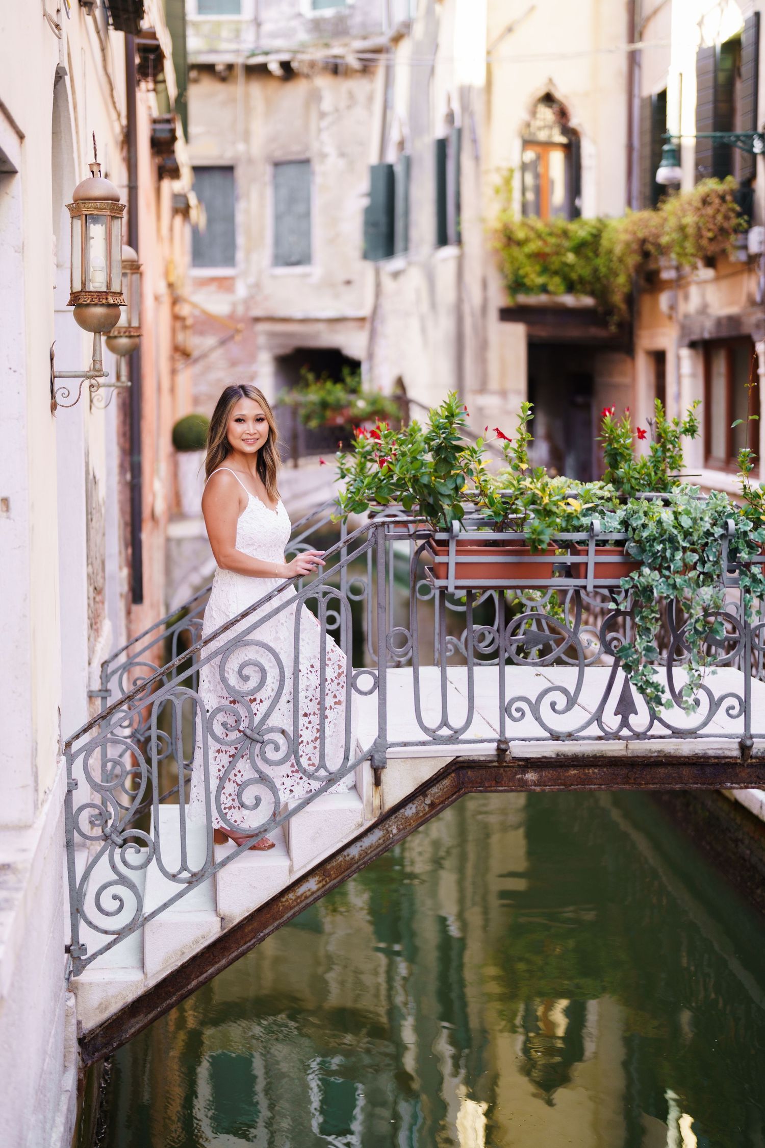 Woman in white dress posing on ornate iron bridge over Venice canal surrounded by lush greenery.