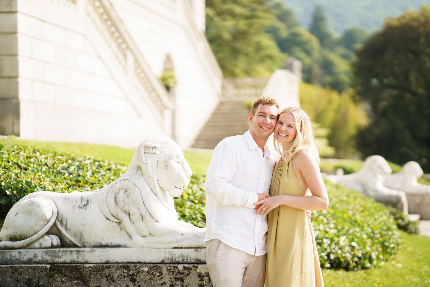 Couple posing beside lion statue at elegant estate with lush gardens, woman in yellow dress, man in white shirt.