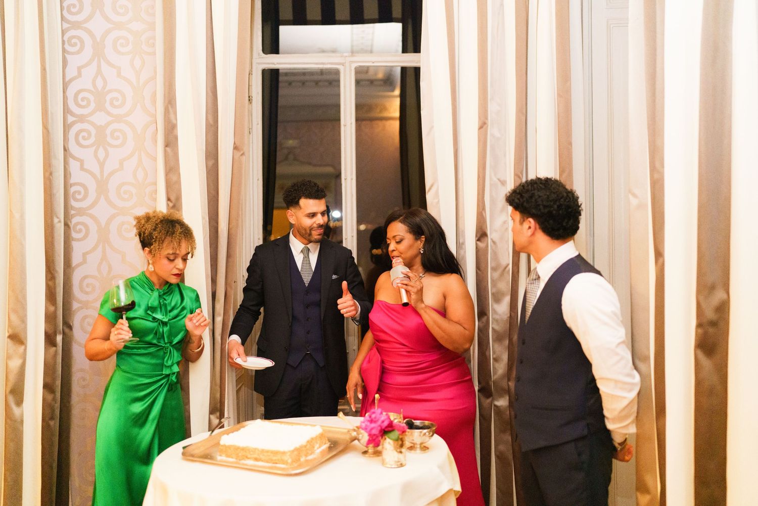 Four elegantly dressed wedding guests gather around a cake table, laughing and celebrating at a formal indoor reception.