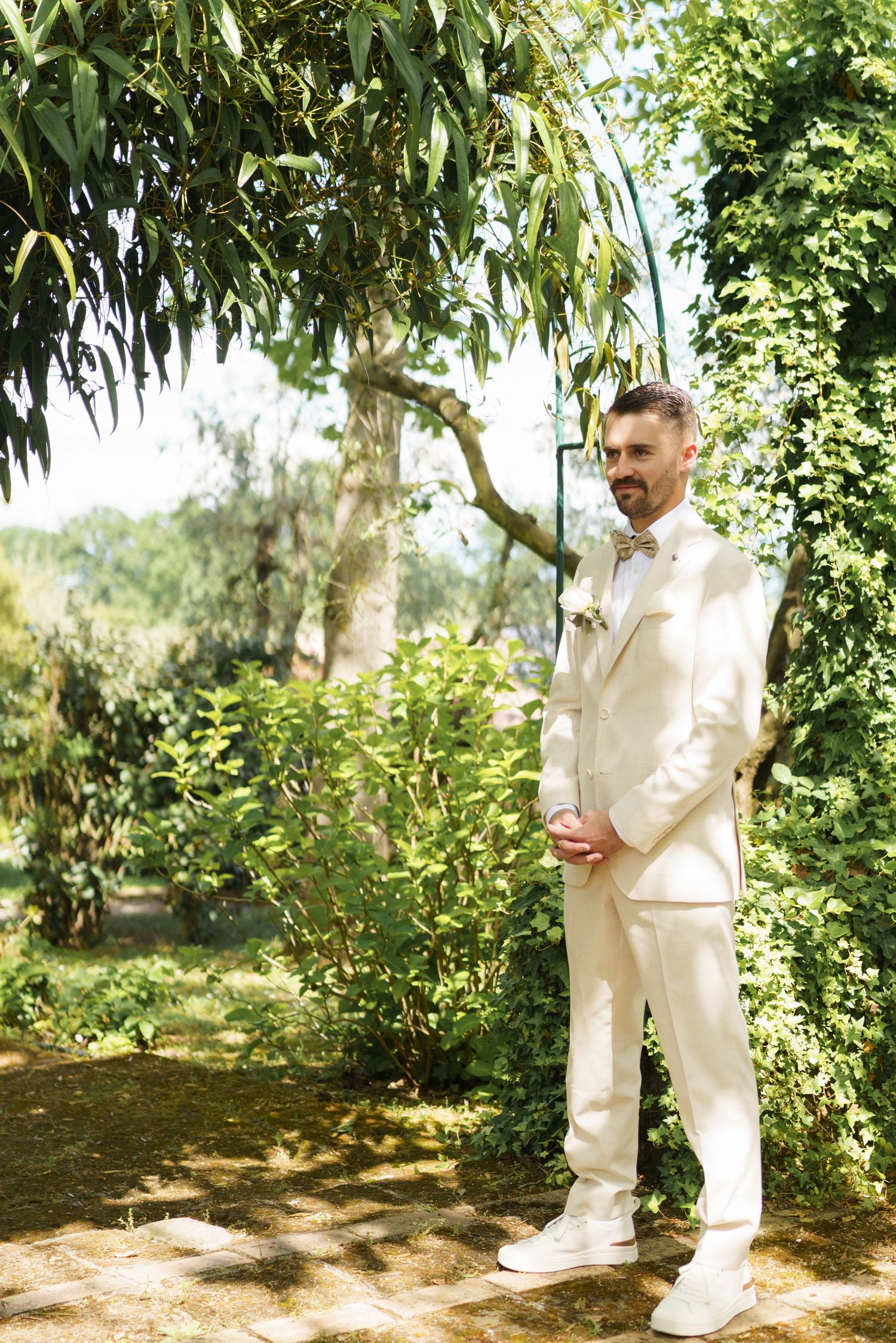 Groom in white suit standing under green archway in sunlit garden on wedding day.