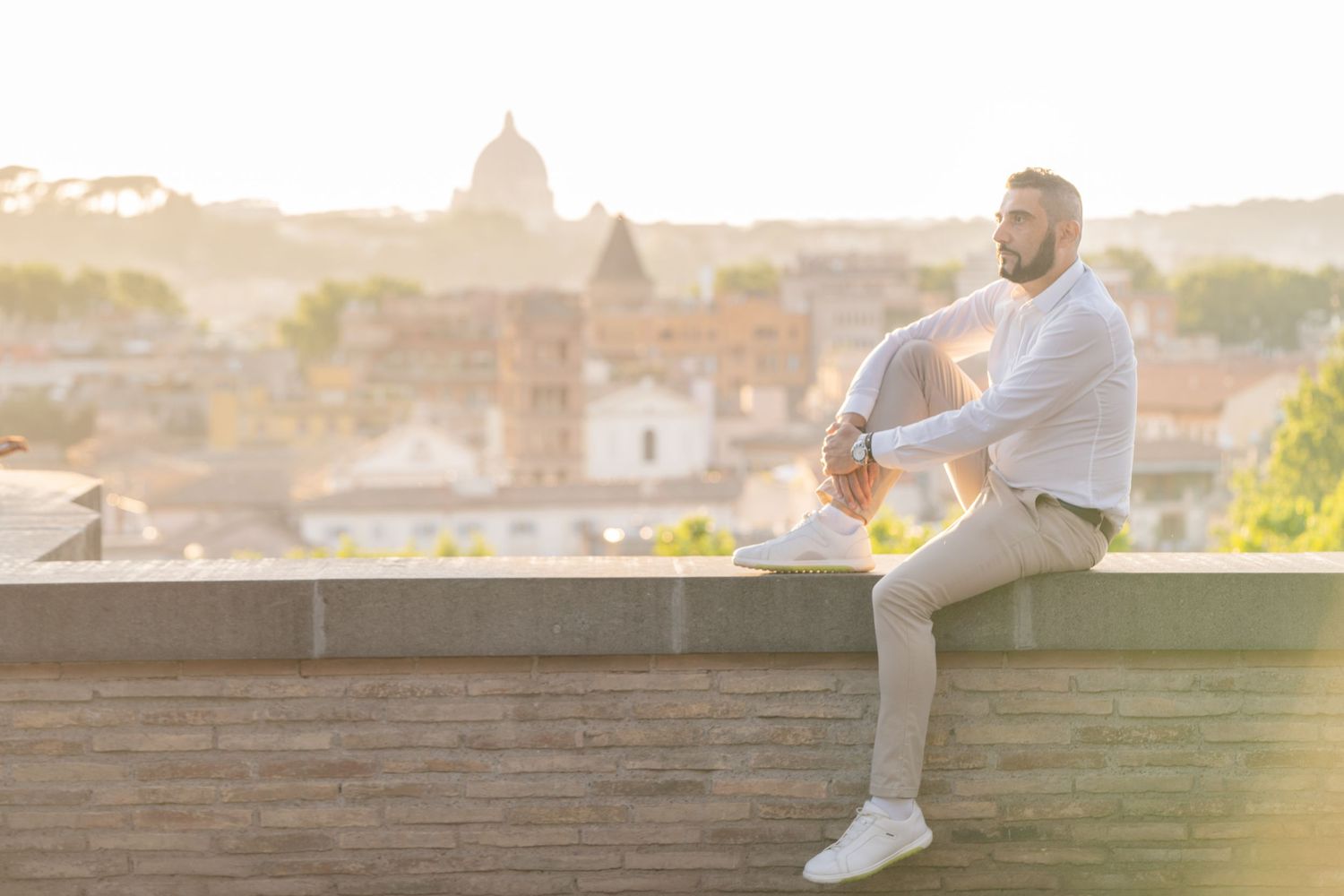 Man sitting on a rooftop wall in Rome, Italy, with a stunning cityscape and golden sunset in the background.