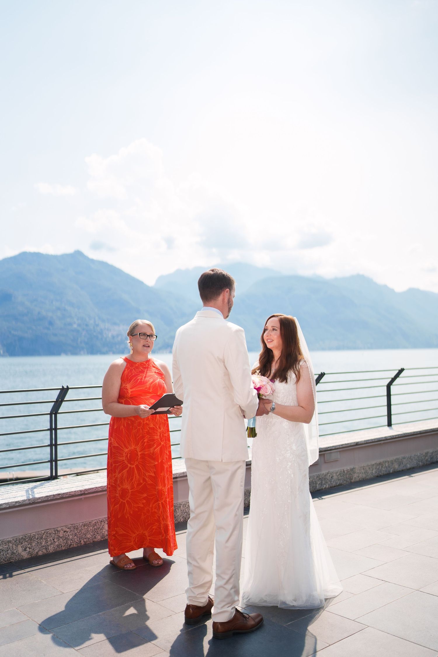 Couple exchanging vows with officiant in red dress, mountain lake backdrop, outdoor wedding ceremony.