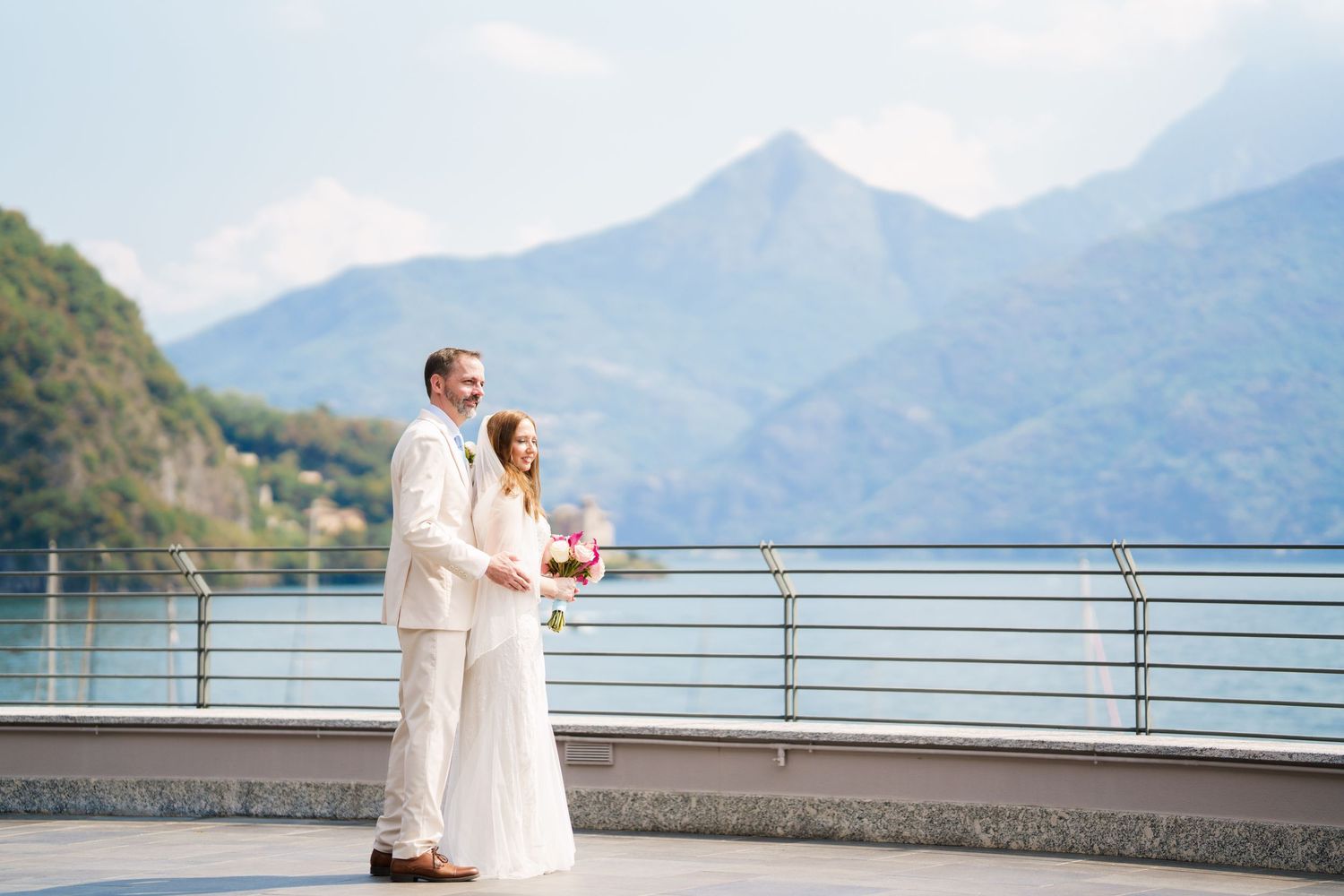Bride and groom stand on a lakeside terrace with stunning mountain backdrop, bride holding pink bouquet.