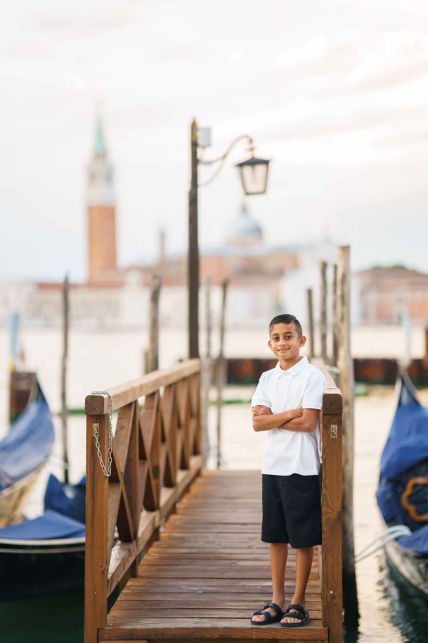 Young boy with arms crossed standing on a wooden dock in Venice, Italy, with gondolas and San Giorgio Maggiore in background.