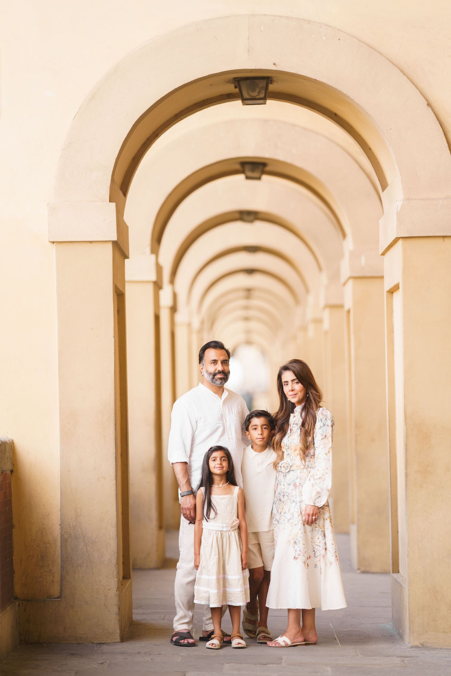 Family of four in white outfits posing under elegant arched colonnade corridor for a portrait photo.
