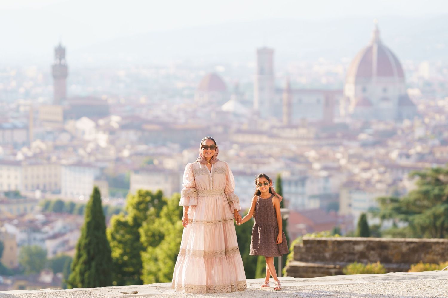 Woman and girl hold hands on Florence overlook with Duomo and Palazzo Vecchio visible in misty skyline.