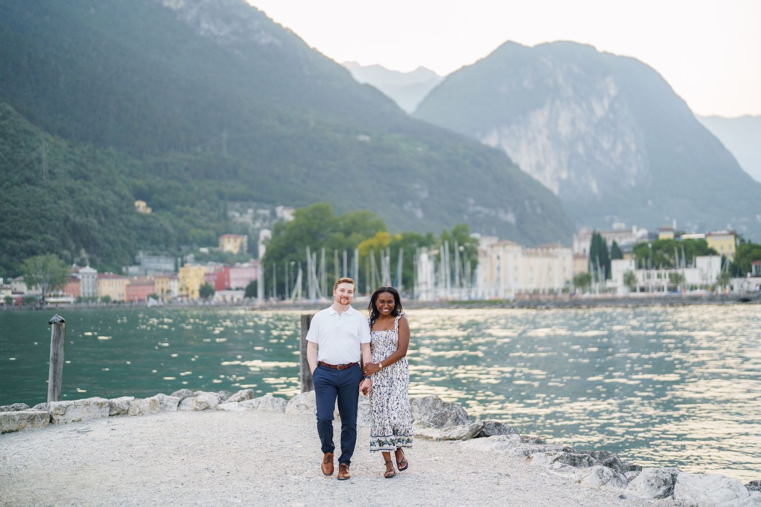 Smiling interracial couple strolling along a scenic lakeside path with colorful town and mountains.