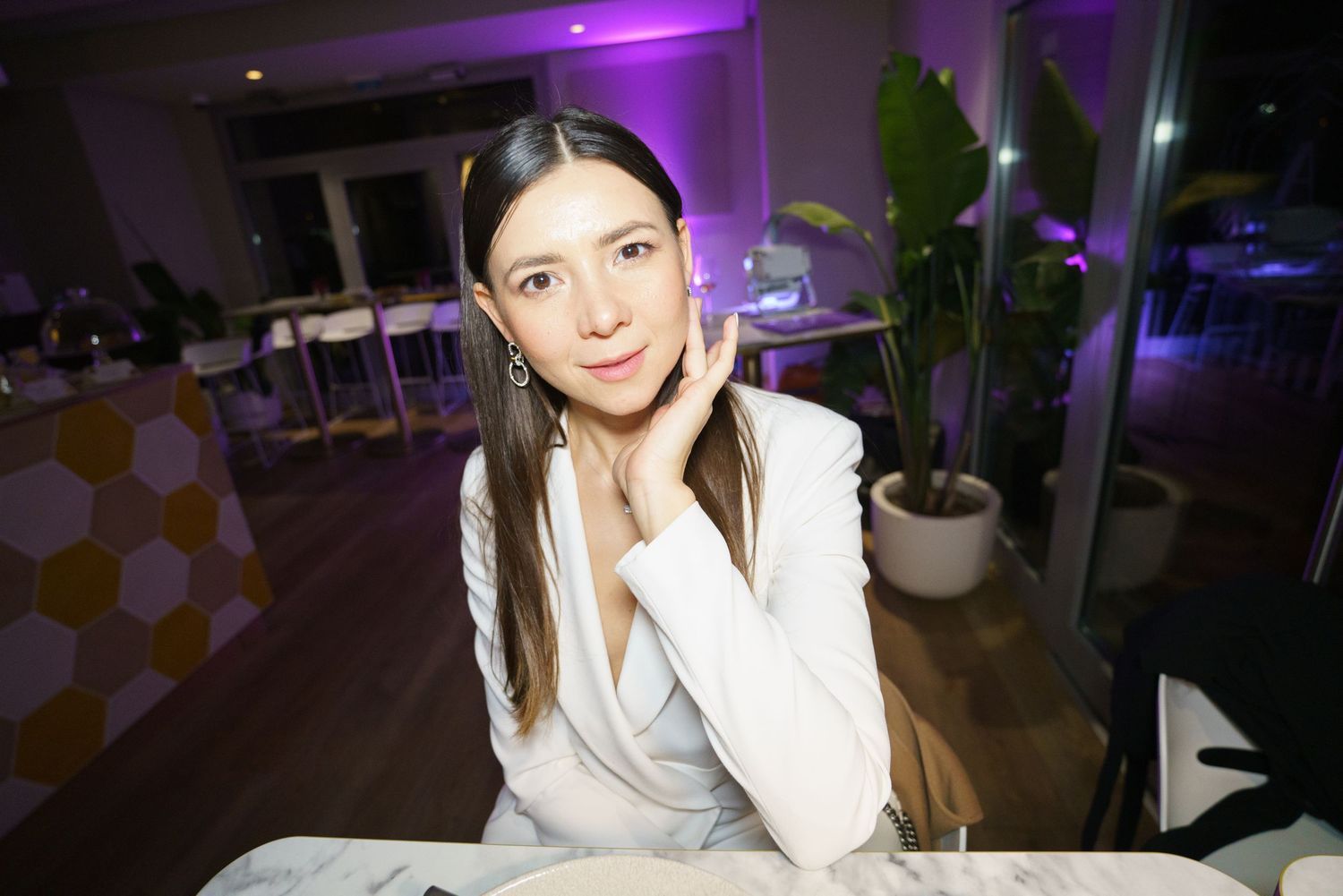 Young woman in white blazer poses elegantly at a restaurant table with purple ambient lighting.