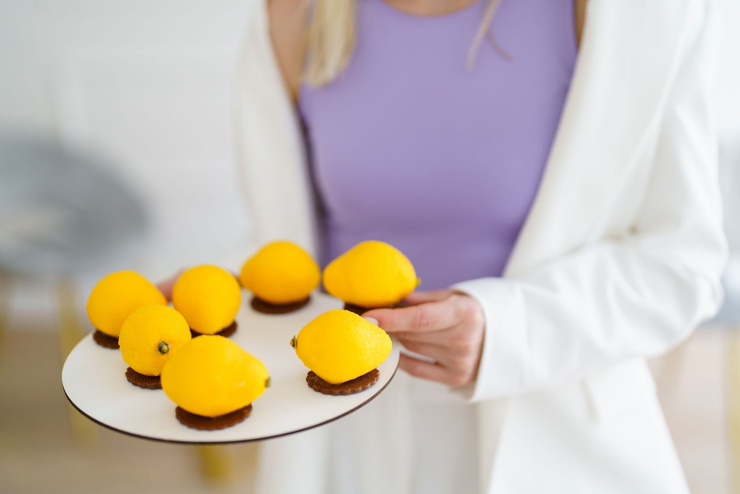 Woman in white blazer holding a plate of elegant lemon-shaped mousse desserts with chocolate bases.