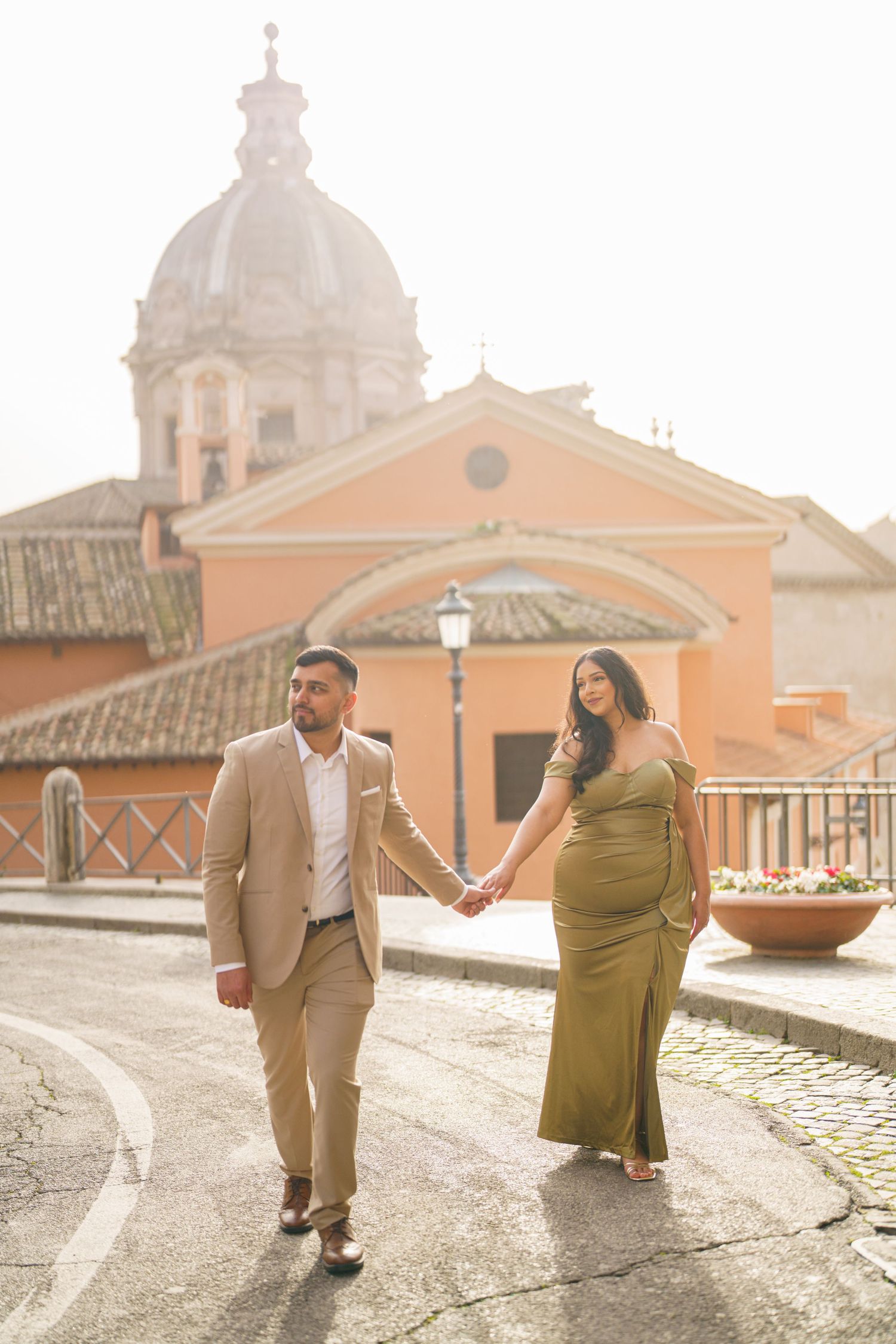 Pregnant couple holding hands walking on cobblestone street with Roman church dome backdrop in golden light.
