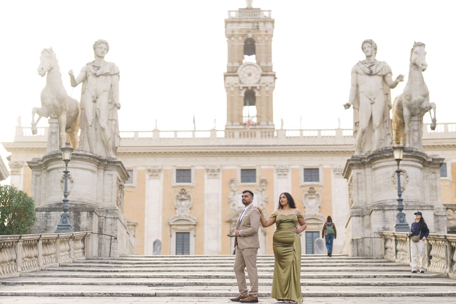 Couple posing on the grand steps of Capitoline Hill in Rome, flanked by classical statues and a historic palazzo.