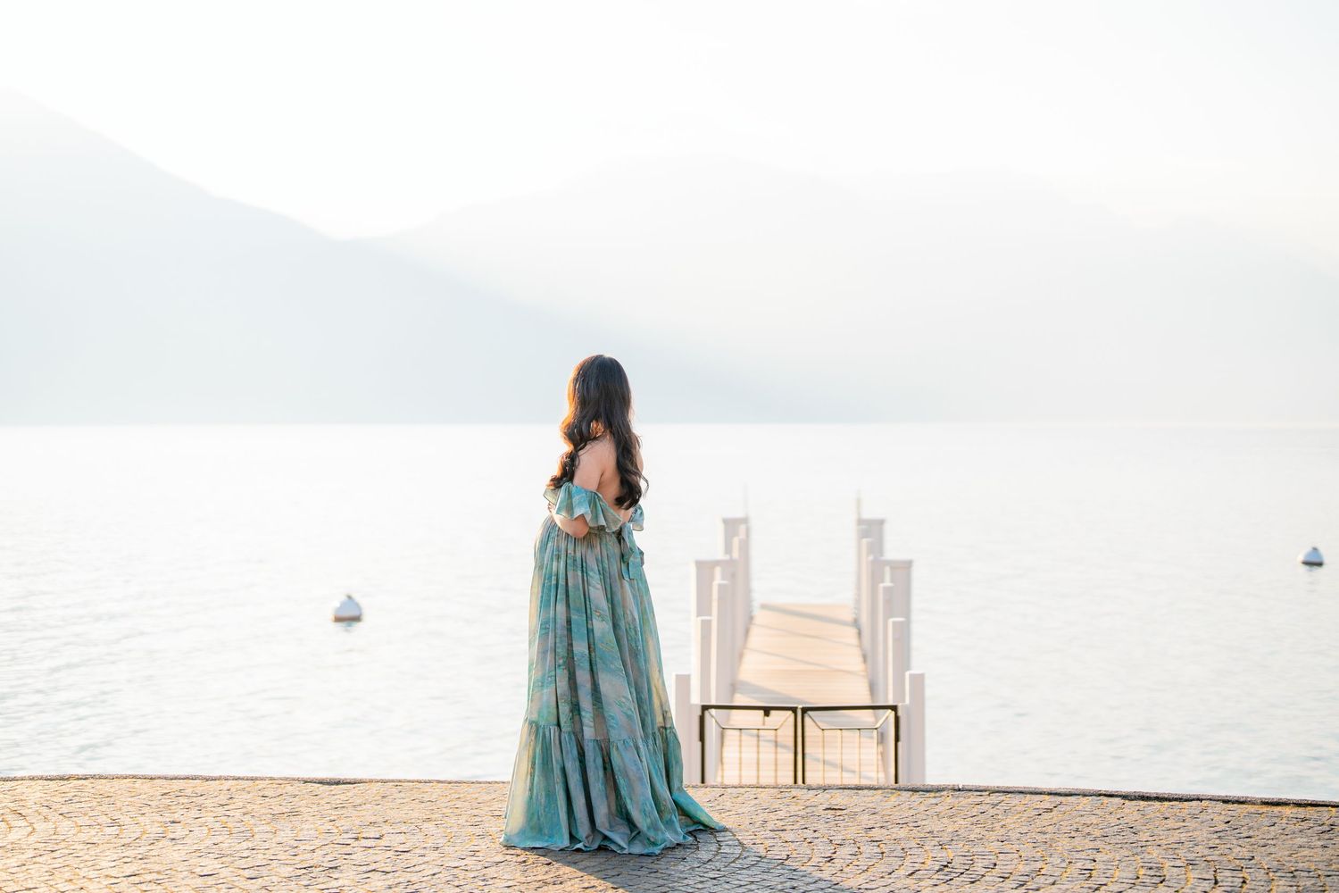 Woman in teal floral gown stands on lakeside pier gazing at misty mountain lake scenery.