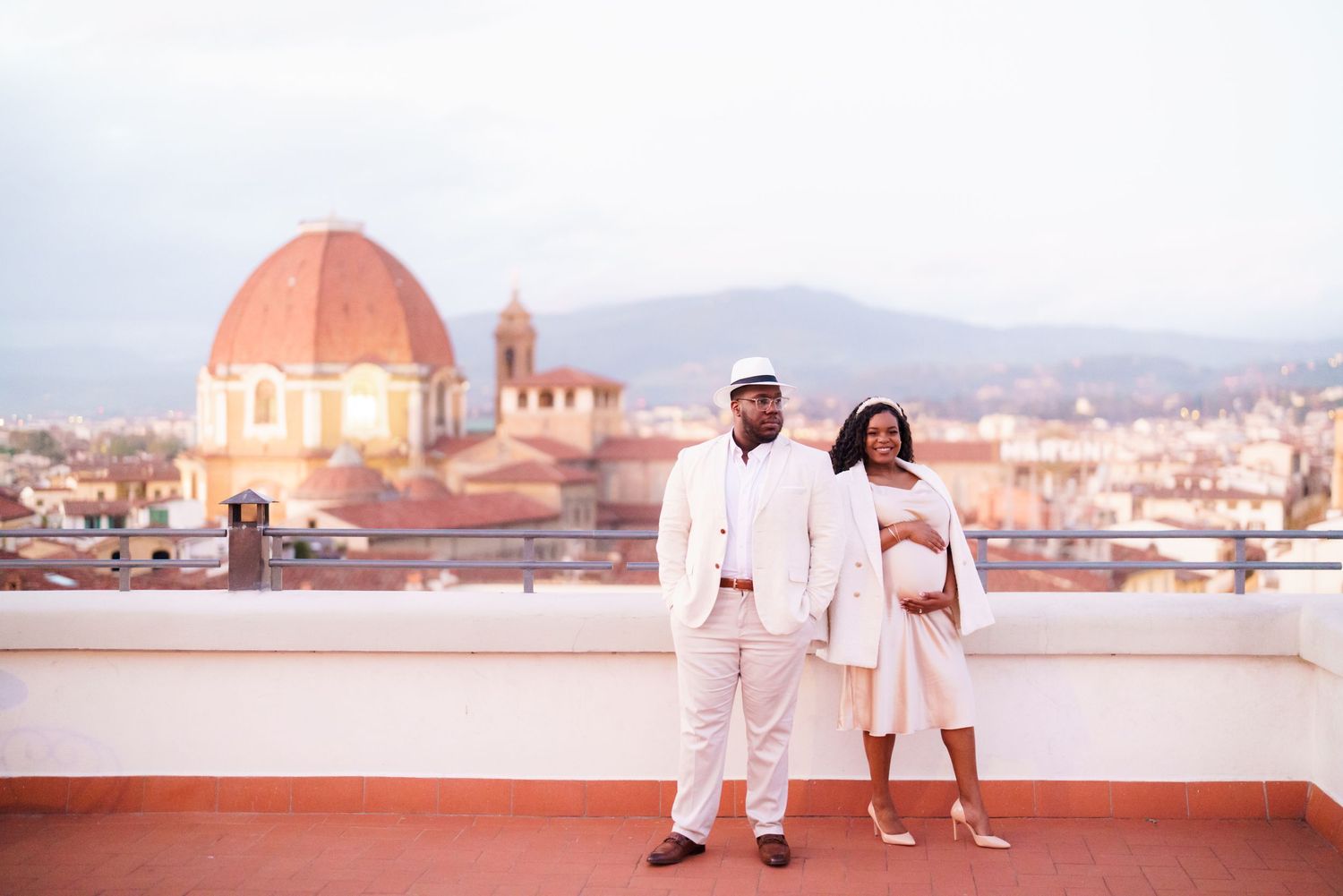 Pregnant couple in white outfits posing on a rooftop terrace with Florence cathedral dome and cityscape behind them.